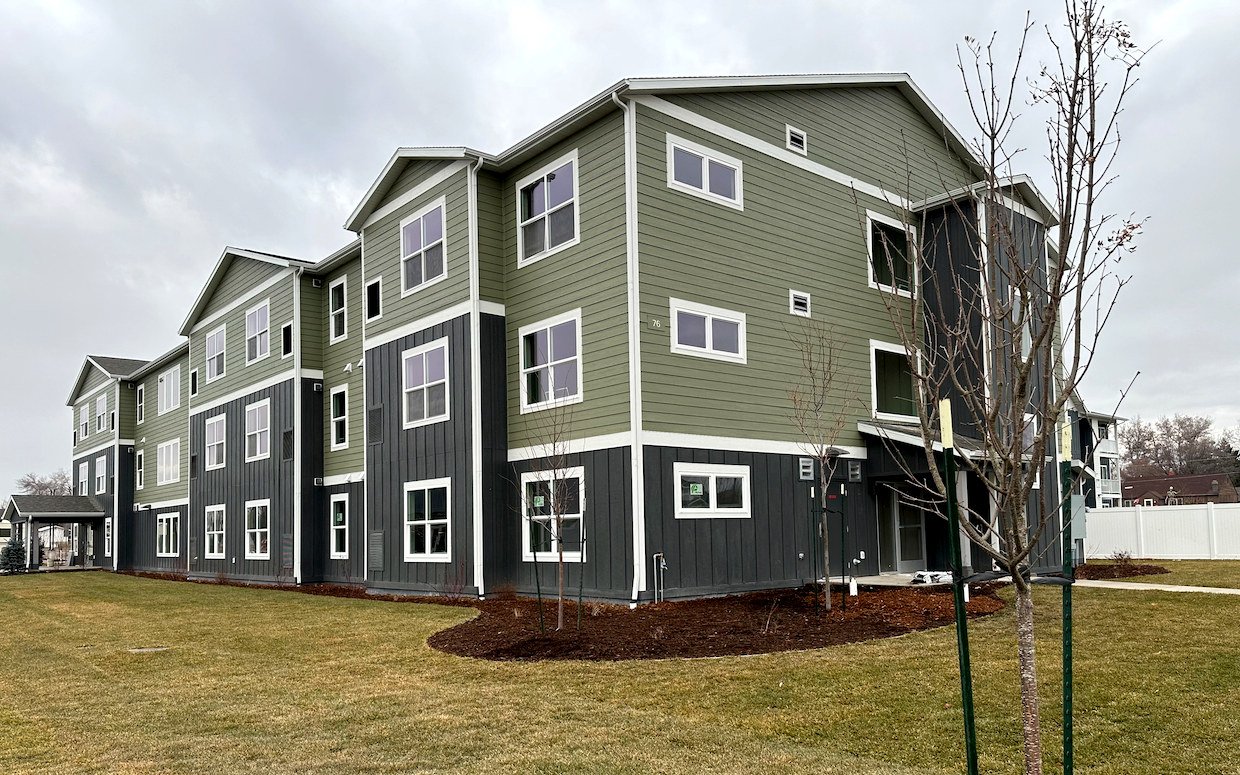 Corner of a three-story apartment building with green and dark gray siding and white trim, with a lawn, fresh mulch beds and young trees under an overcast sky.