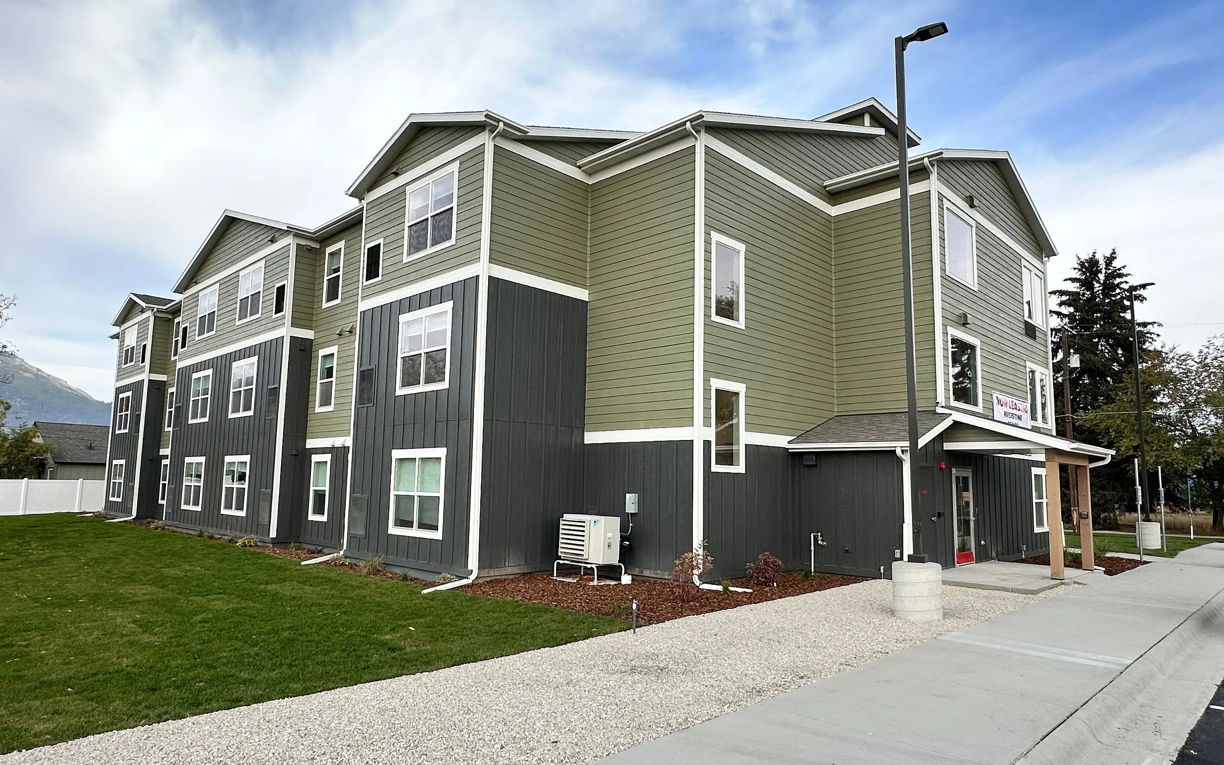 Side of a three-story apartment building with green and dark gray siding, a lawn, a gravel path and a covered entrance, under a partly cloudy sky.