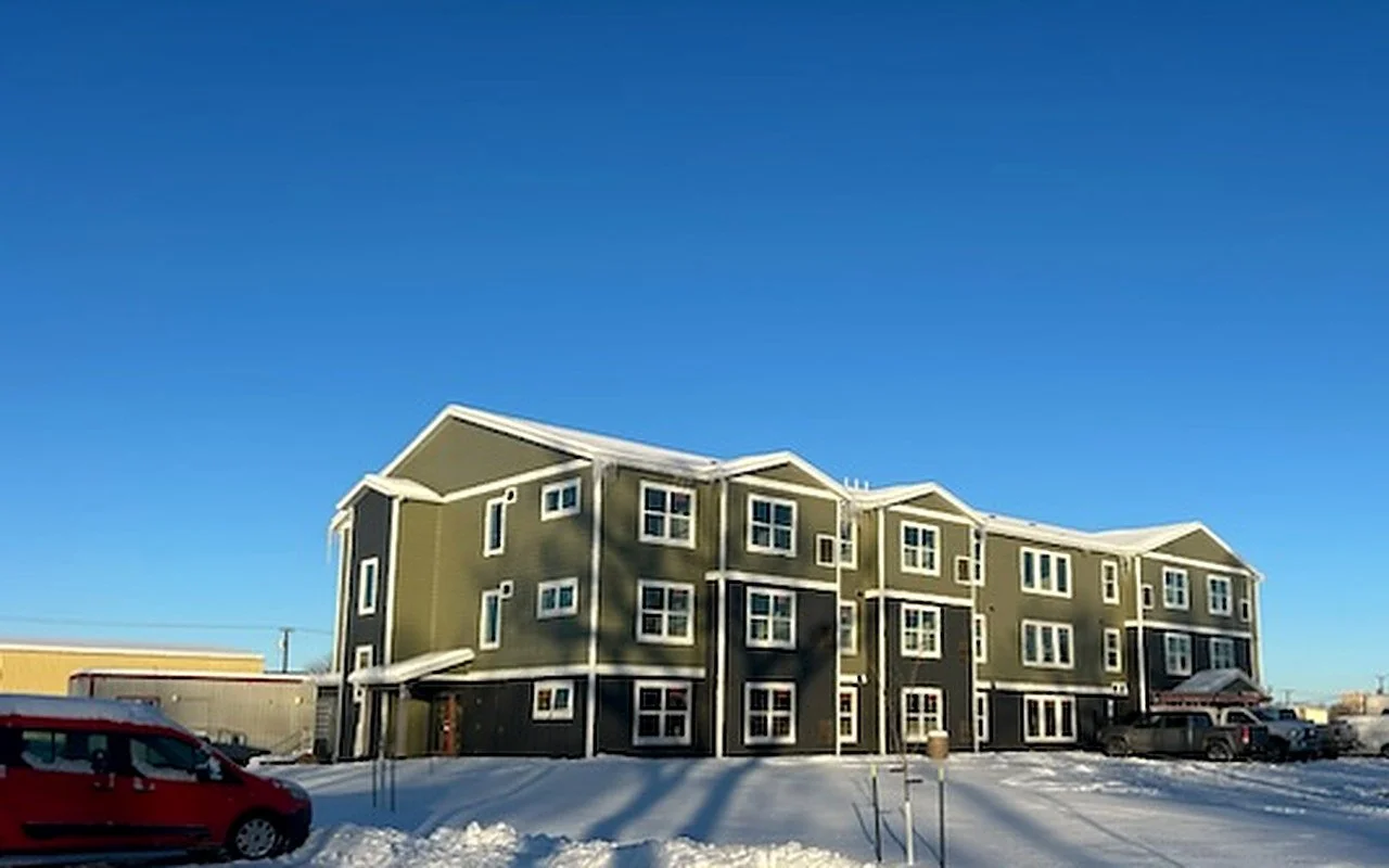 Three-story apartment building with green and dark gray siding and white trim, seen across a snow-covered lot under a deep blue sky.