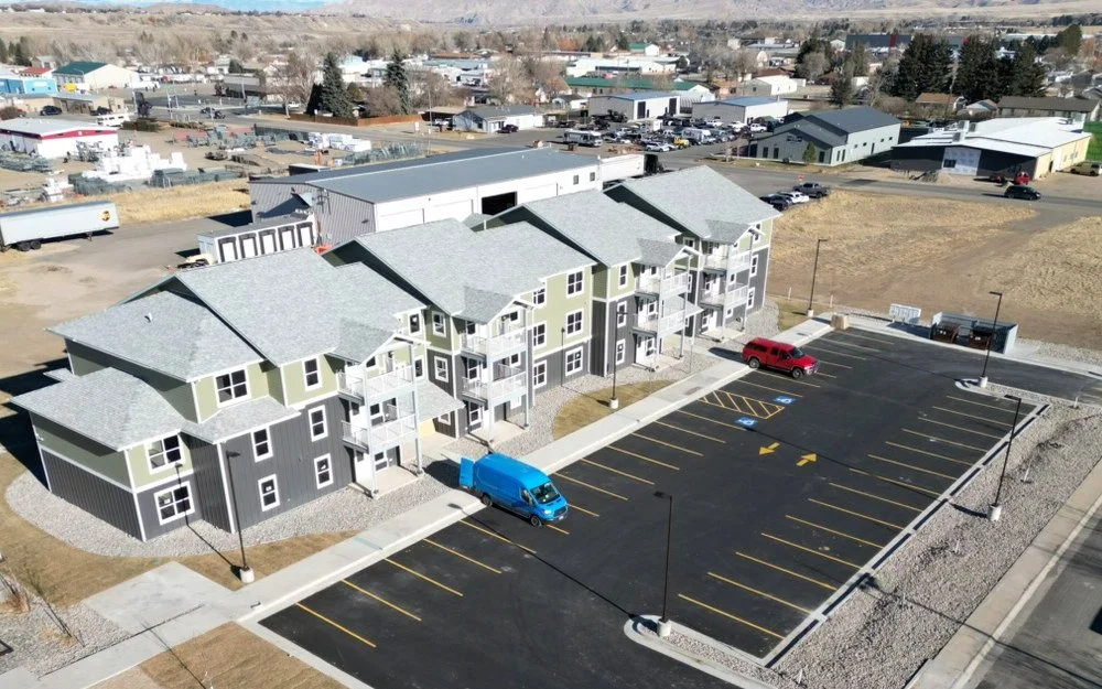 Aerial view of a three-story apartment building with green and gray siding beside a large parking lot, with the town of Cody in the background.