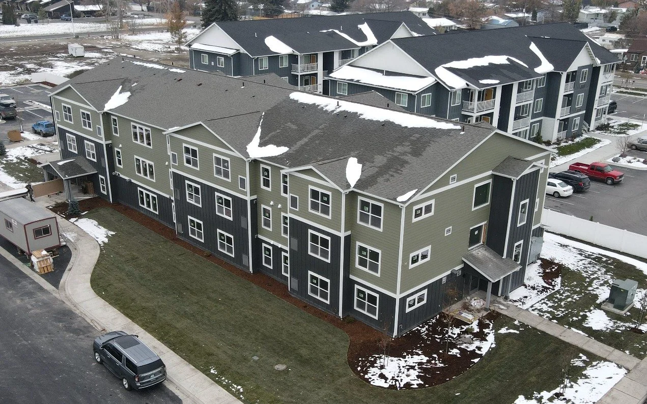 Aerial view of a three-story apartment building with green and dark gray siding and a curving lawn, with light snow on the roofs and ground and neighboring buildings behind.