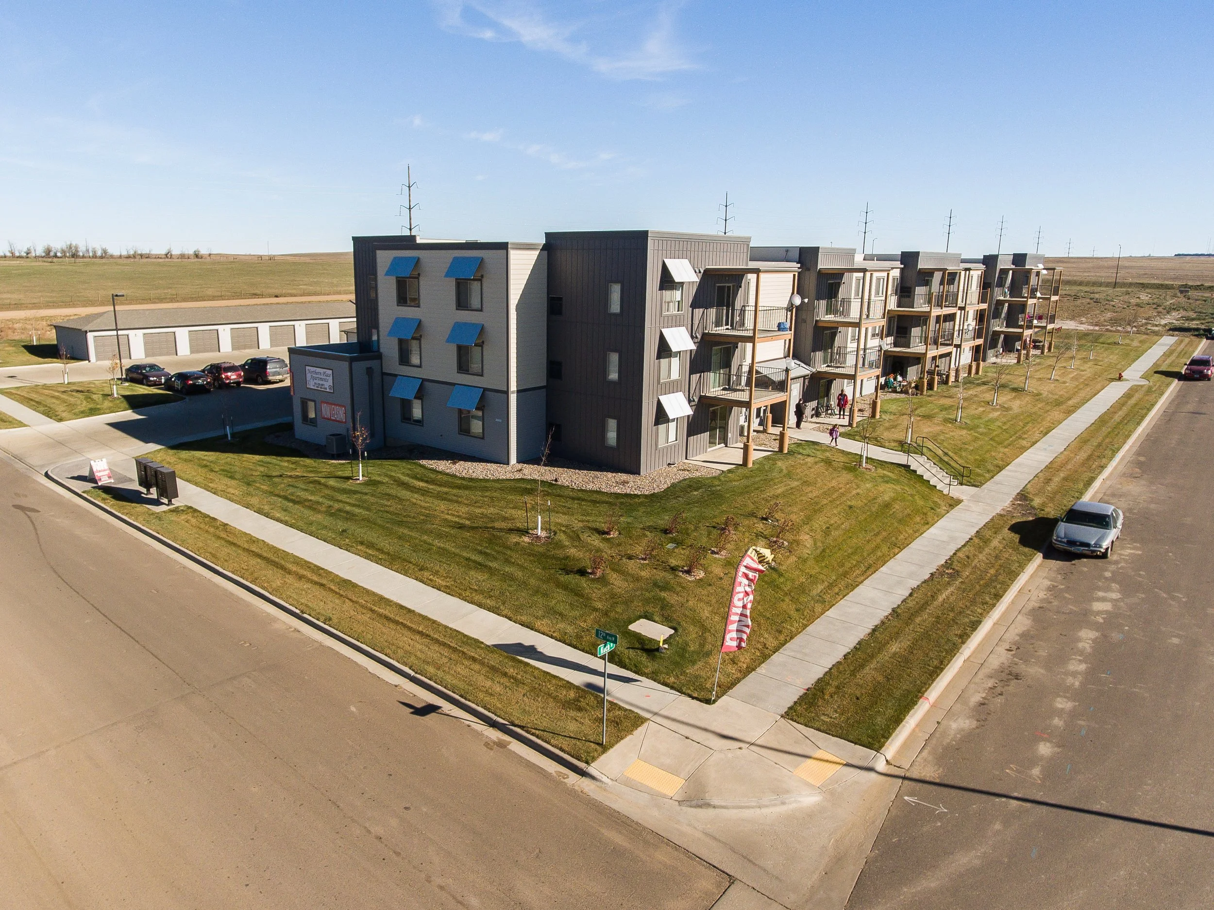 Aerial view of a multi-story apartment complex with walking paths, parking lot, and grassy areas, in a suburban setting under a clear blue sky.