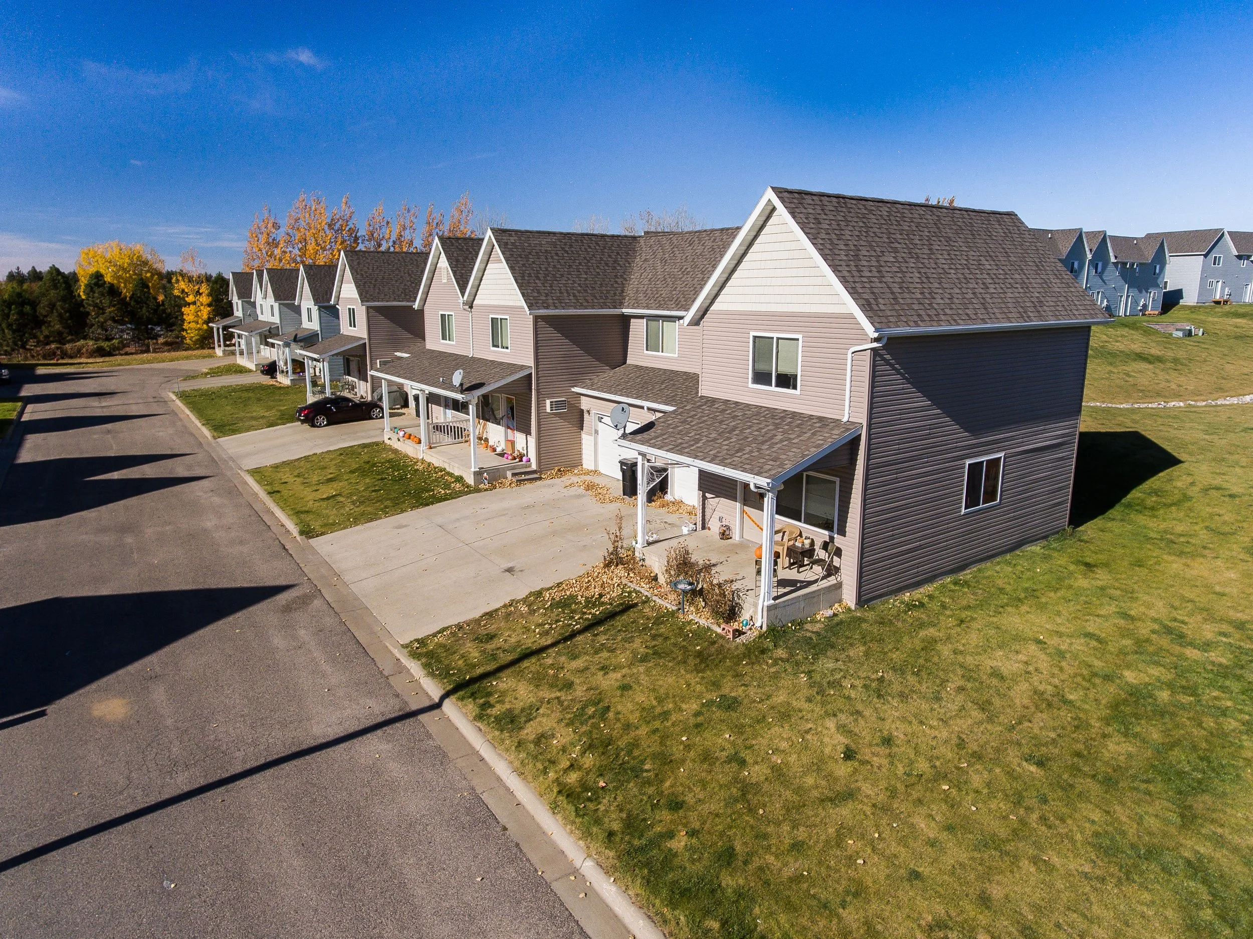 Aerial view of a row of townhouses with small front yards and driveways, some with parked cars, under a clear blue sky.