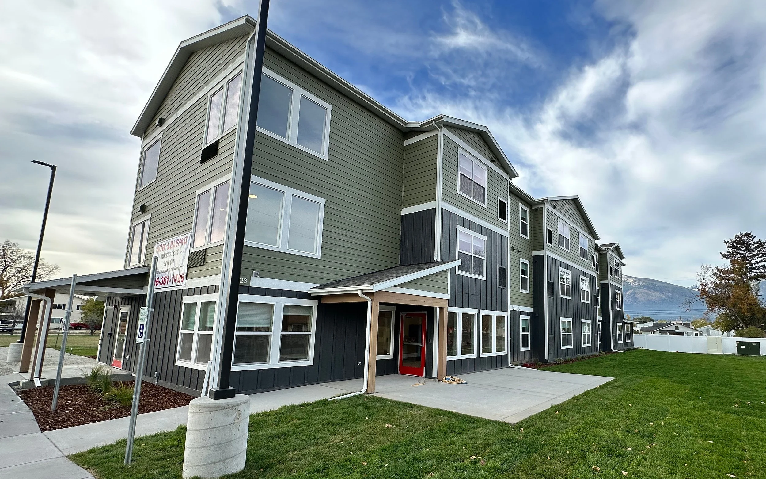 Corner of a three-story apartment building with green and dark gray siding, a covered entrance with a red door and a lawn, with mountains and a cloudy sky in the background.