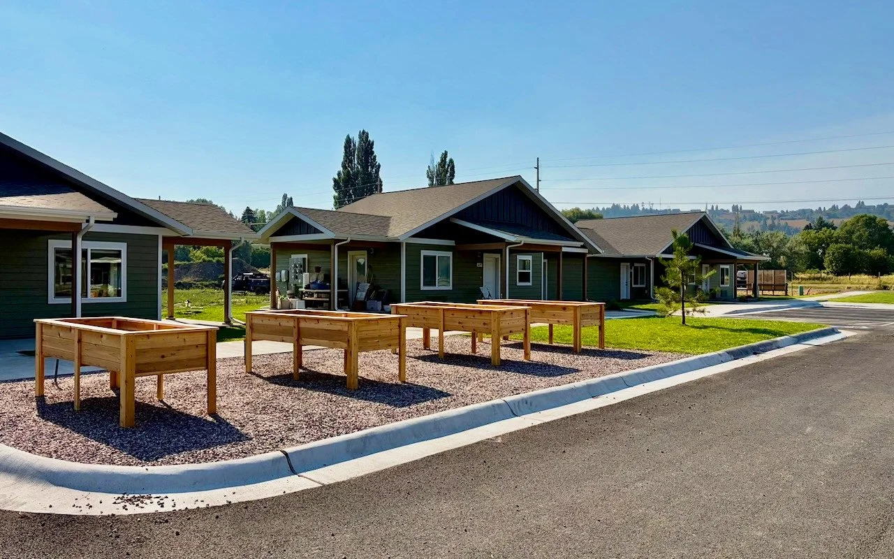 Cedar raised garden beds on a gravel bed in front of single-story homes with green siding and covered porches, under a clear blue sky.