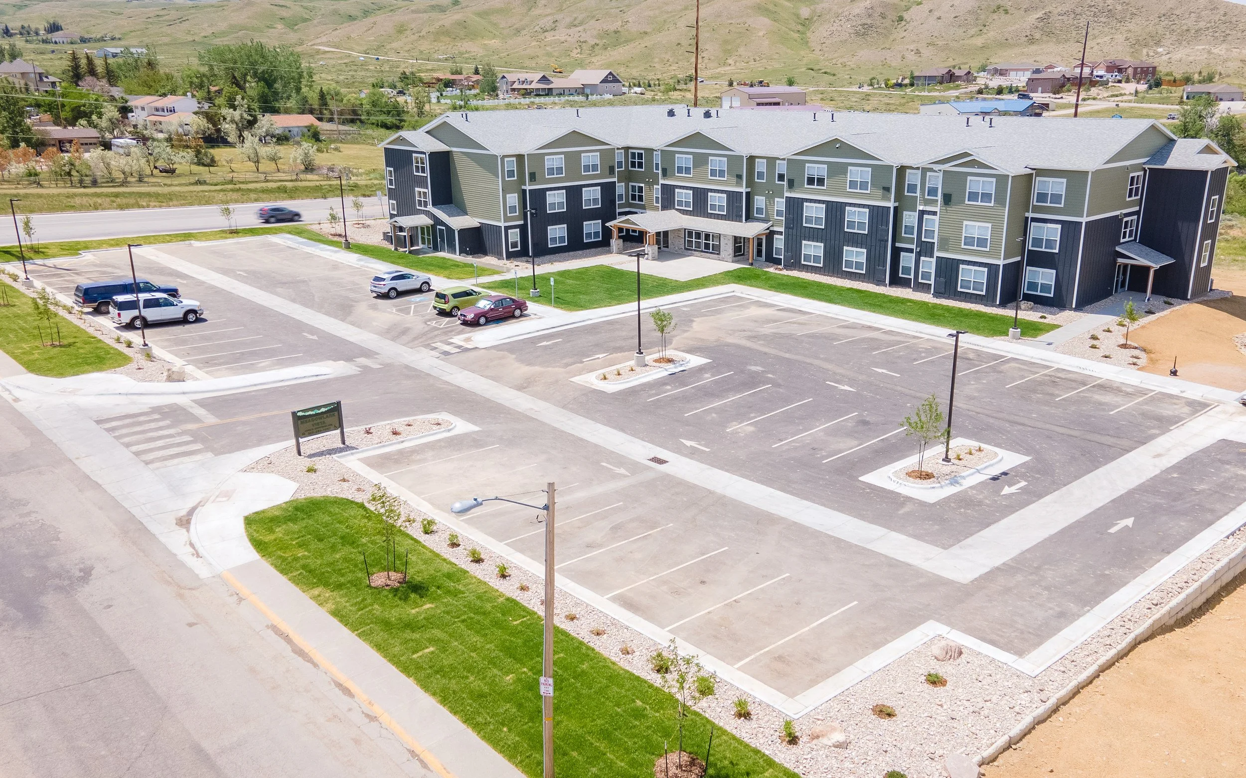Aerial view of a three-story apartment building with a large parking lot, young trees and lawns in front, and grassy hills and homes in the background.