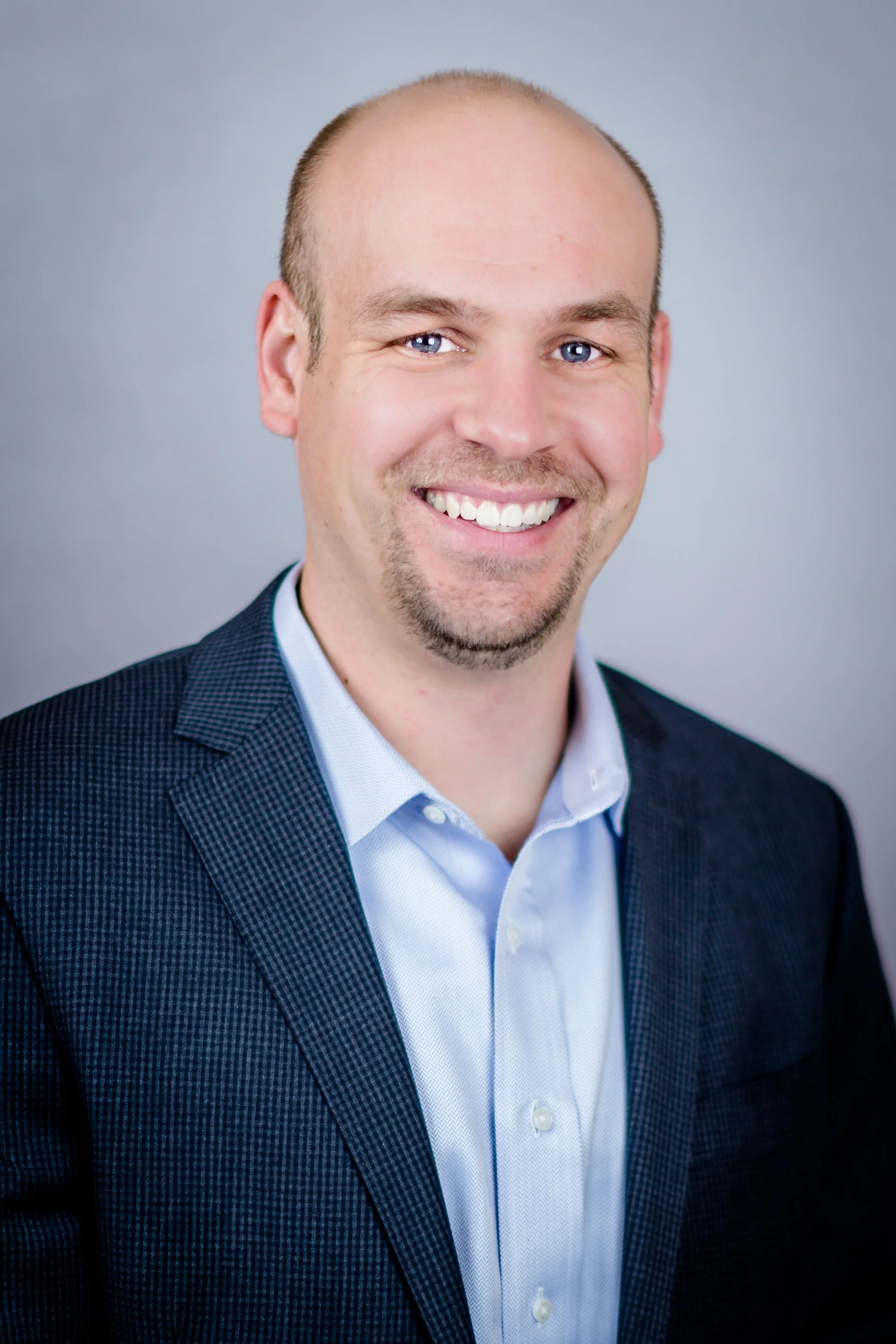 Headshot of a smiling man with short brown hair, blue eyes, wearing a light blue collared shirt and a dark checkered blazer, against a gray background.