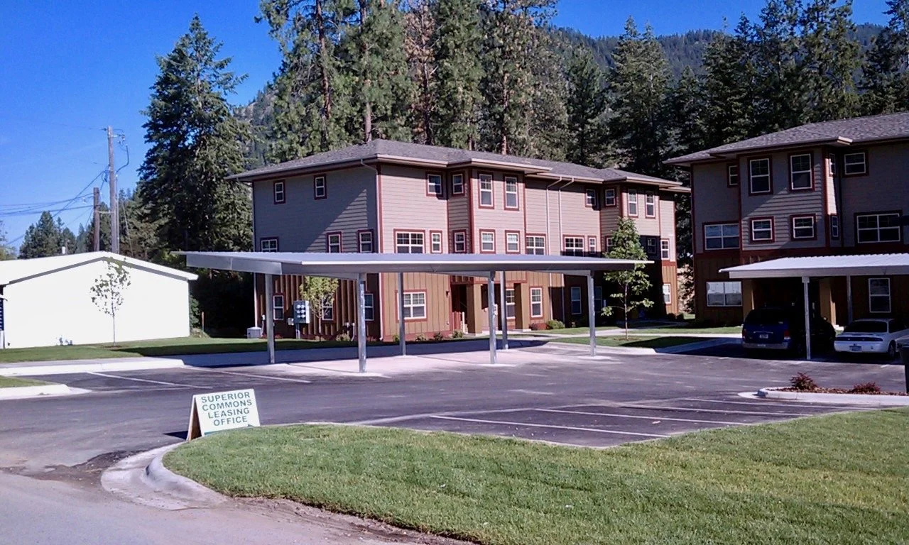 Apartment complex with parking lot, covered carports, and a sign for Superior Commons Leasing Office, surrounded by trees and under a blue sky.