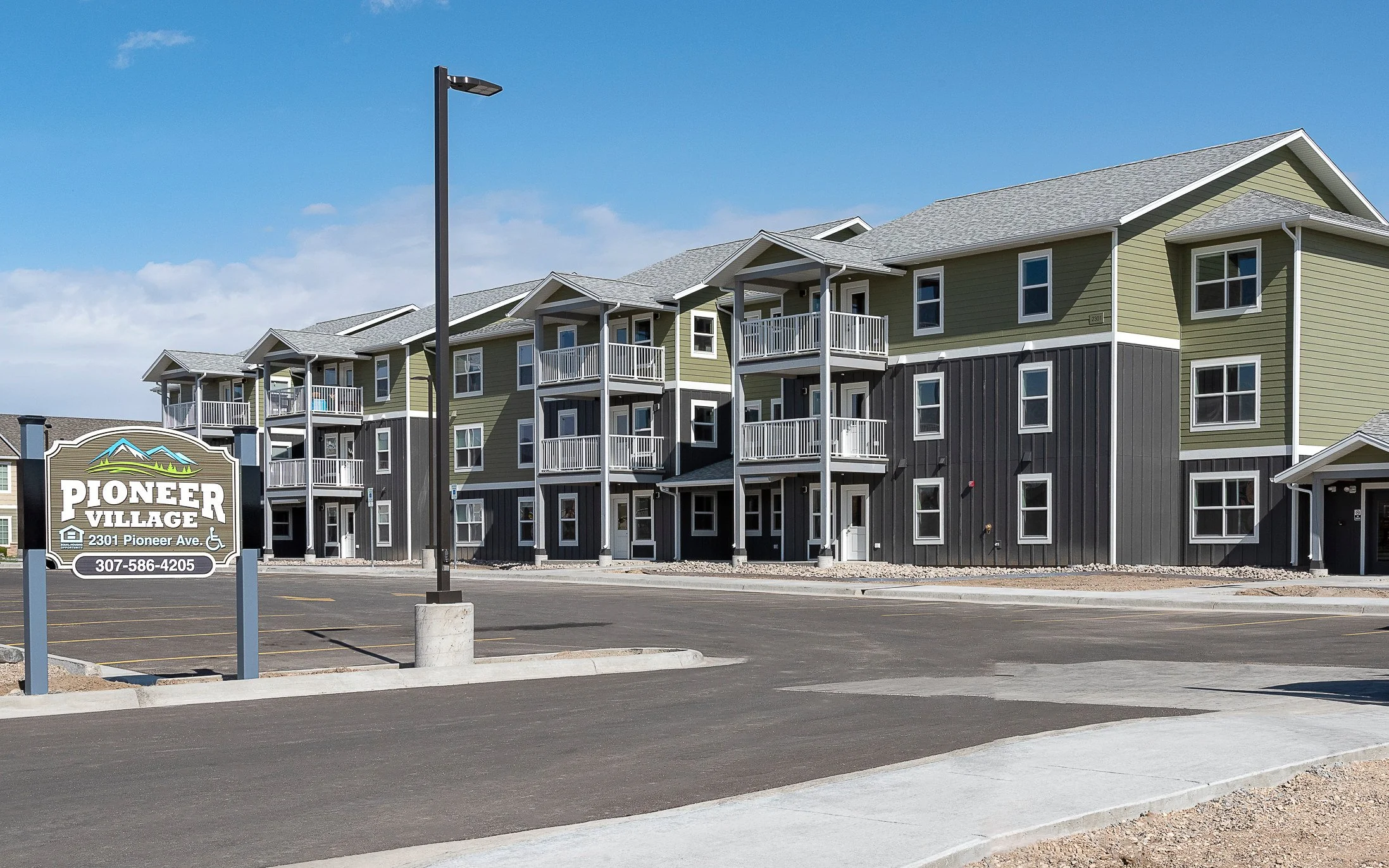 Three-story apartment building with green and dark gray siding and white balconies, with a parking lot and a Pioneer Village monument sign in the foreground under a blue sky.