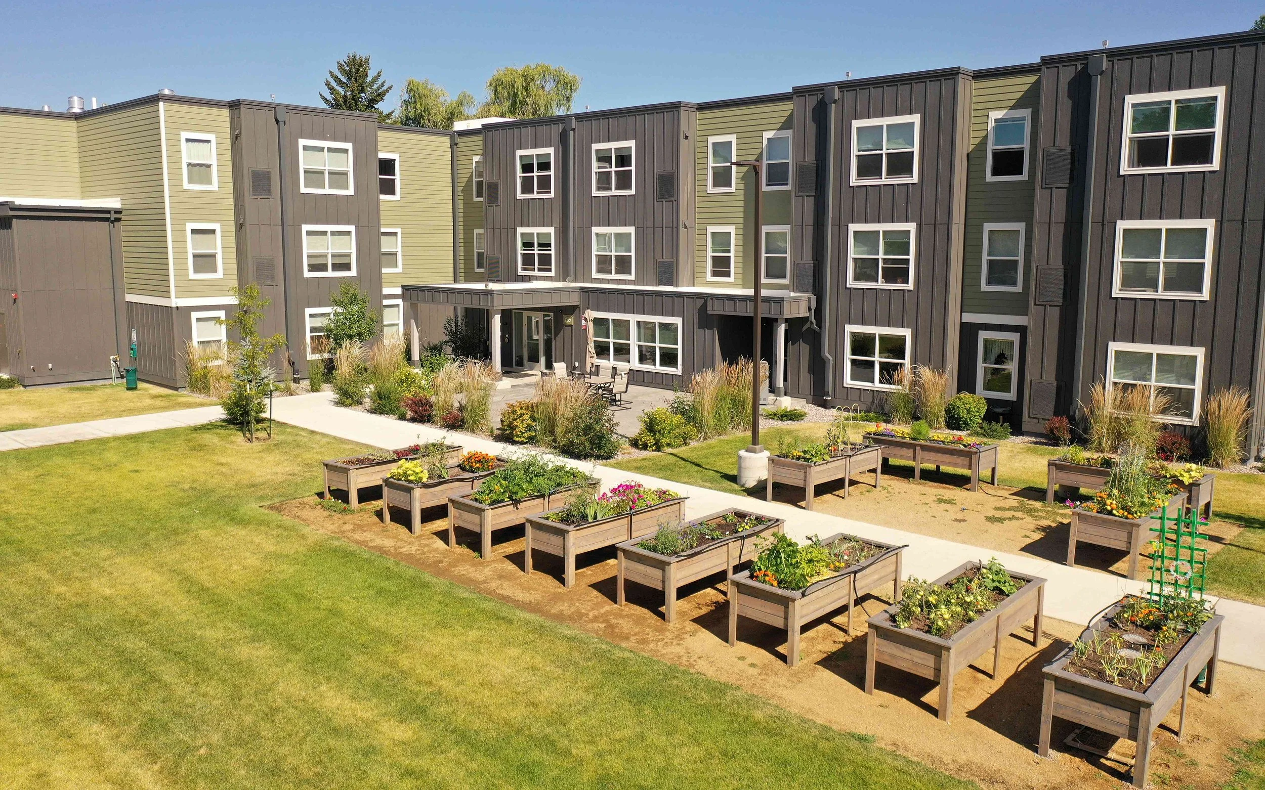 Aerial view of a courtyard with rows of raised wooden garden beds, a lawn and a patio in front of a three-story apartment building with green and dark gray siding.