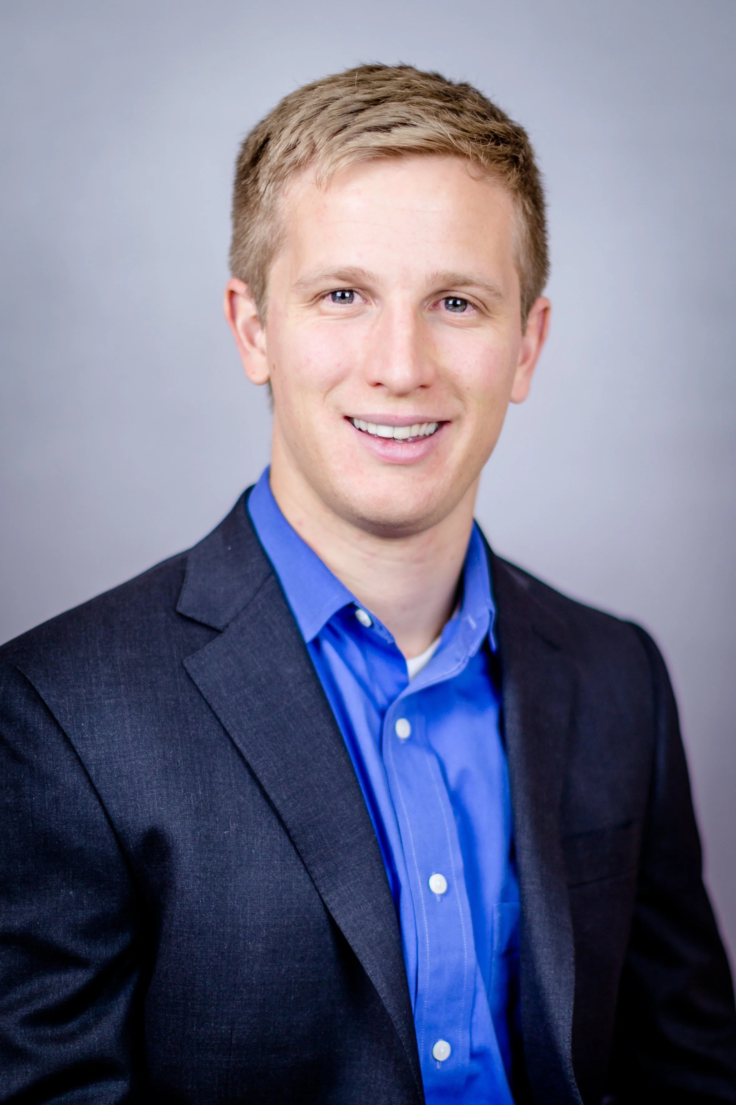 Professional headshot of a young man with blonde hair, blue eyes, wearing a dark blazer and a blue collared shirt, smiling at the camera against a neutral background.