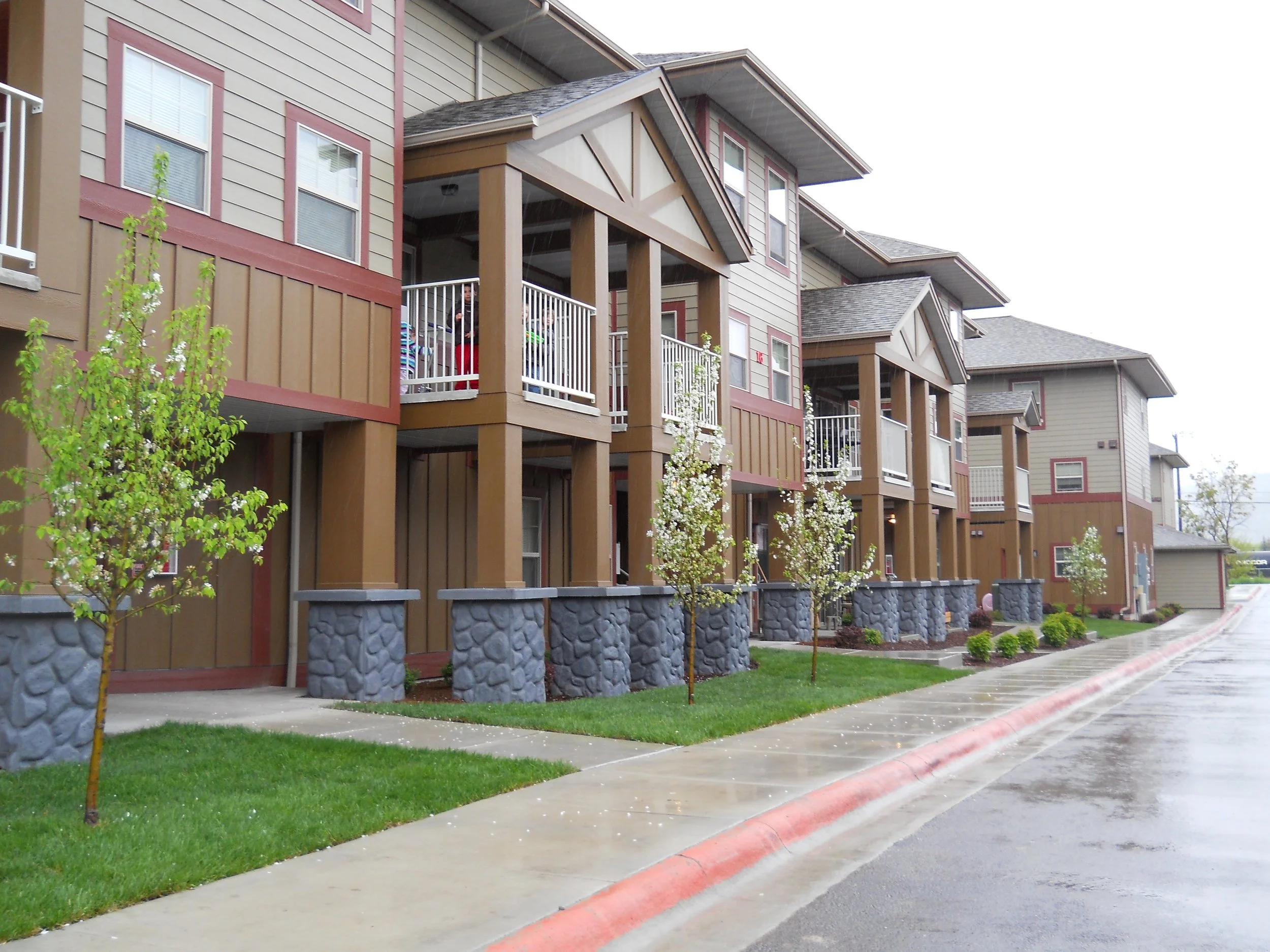 Apartment complex with stone pillars, small trees, and a wet sidewalk.