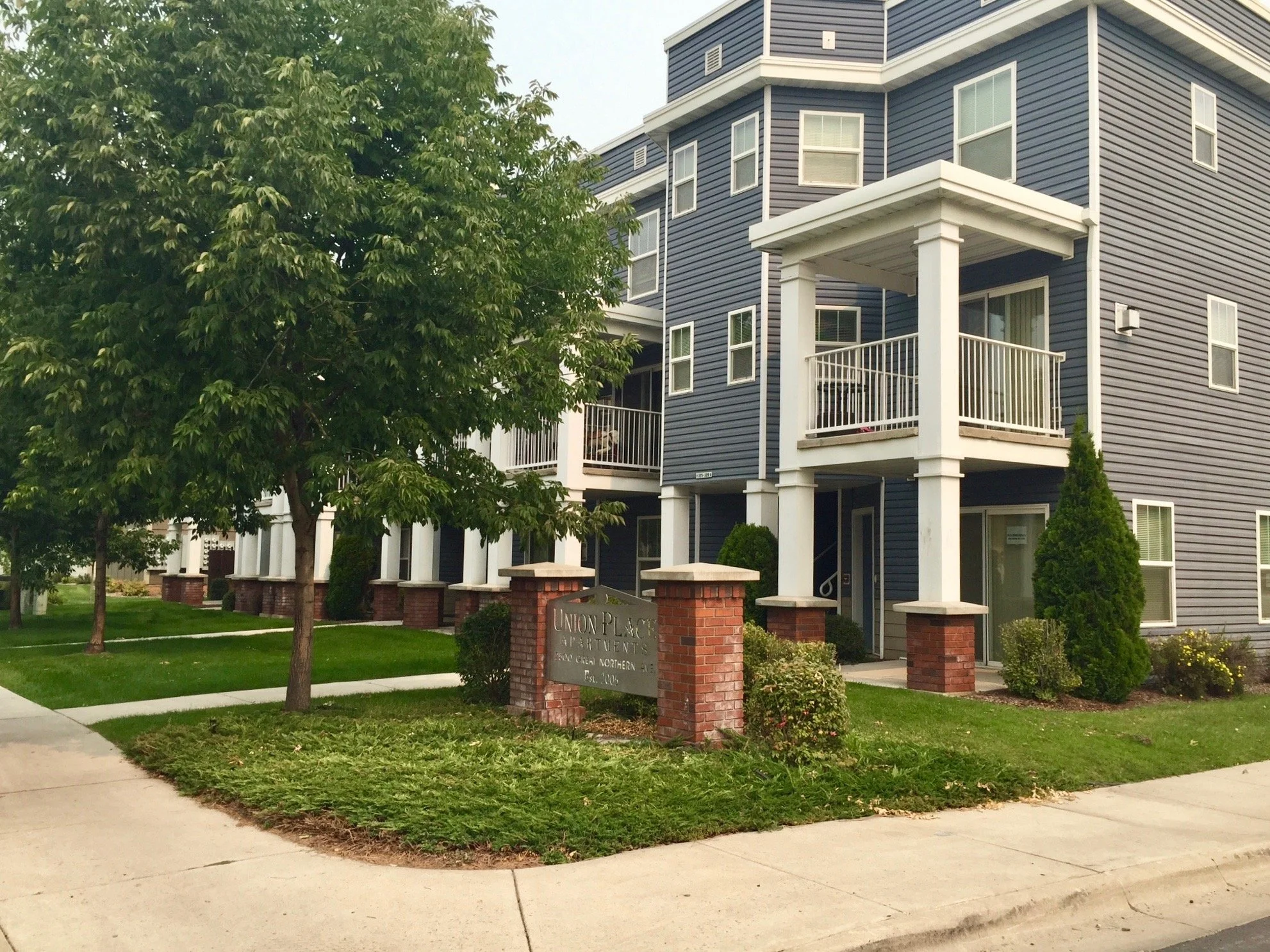 Multi-story apartment building with a blue exterior, white railings on balconies, surrounded by trees and manicured lawns, with a brick sign reading 'Union Place Apartments' near the entrance.