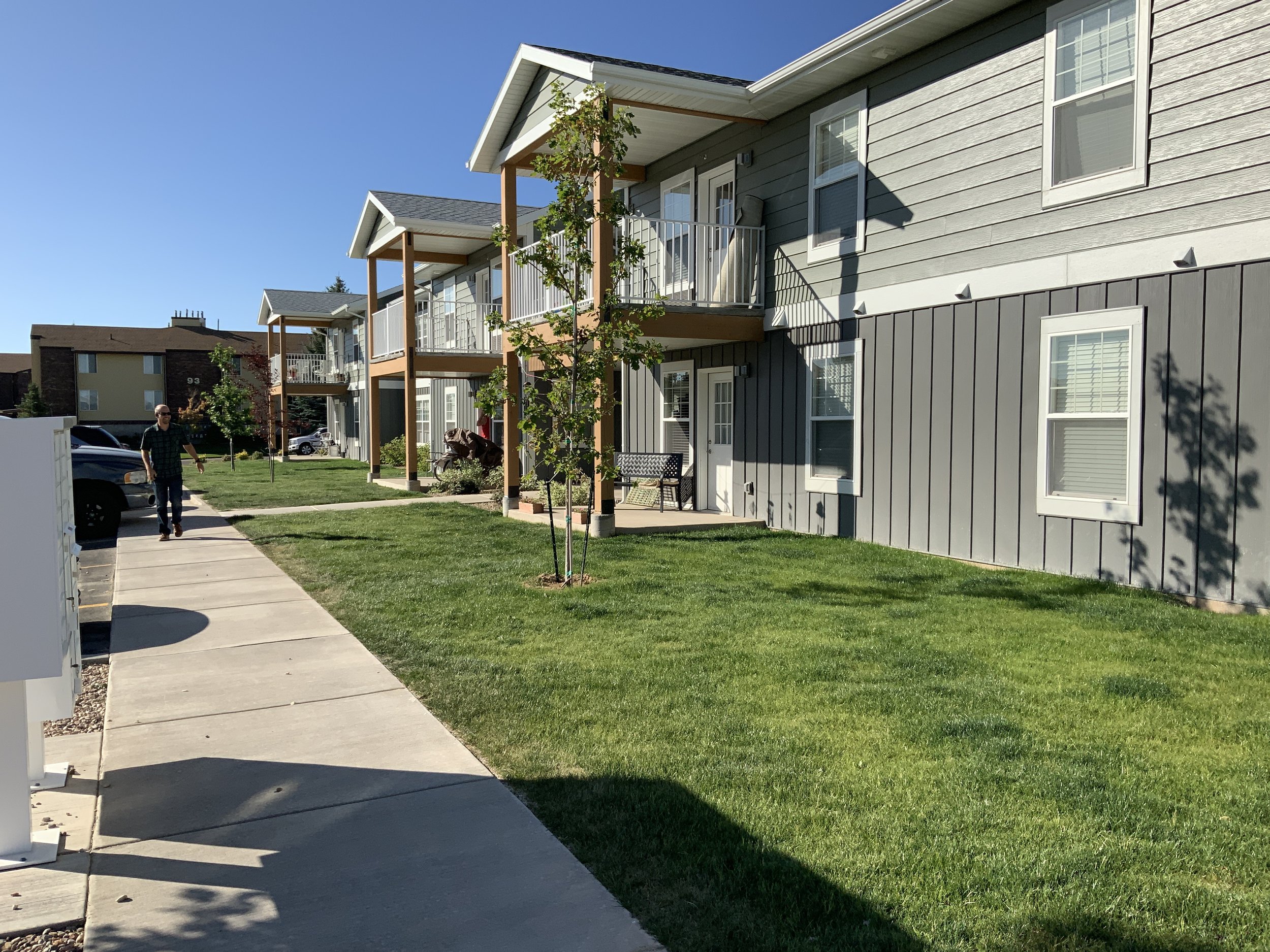 Apartment complex with gray siding, balconies on the second floor, green lawn, sidewalk, small trees, and a man walking.