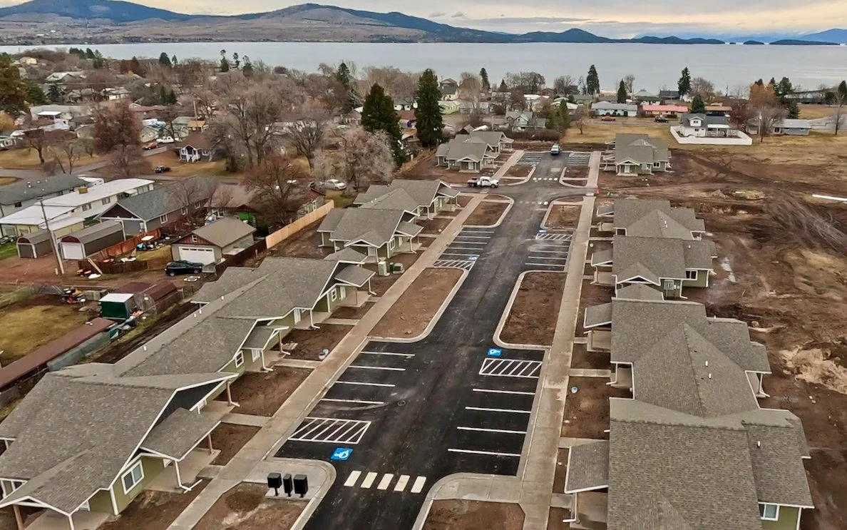 Aerial view of single-story senior fourplexes with gray roofs along a new parking lot, with the town of Polson, Flathead Lake and mountains in the background.