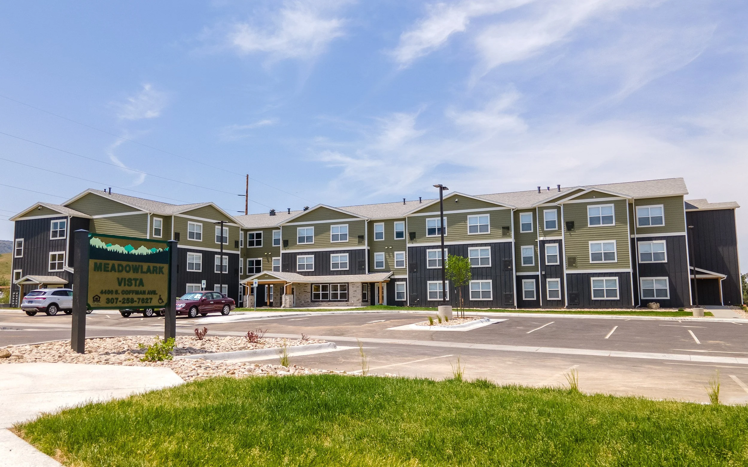 Three-story apartment building with green and dark gray siding behind a parking lot, a lawn and a Meadowlark Vista monument sign, under a blue sky with light clouds.