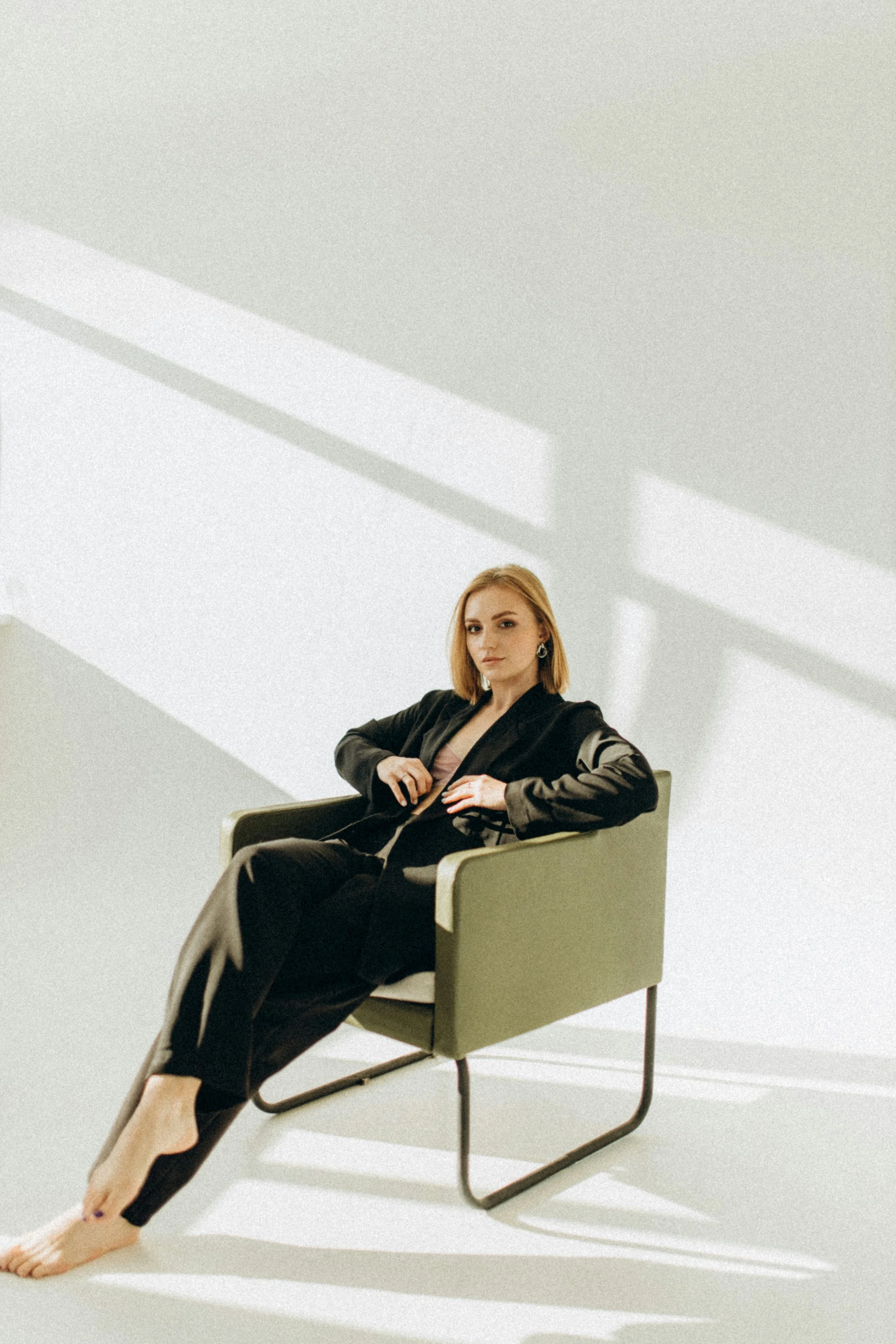 A woman with blonde hair in a black suit sitting in a green chair, with shadows cast on a white wall behind her.