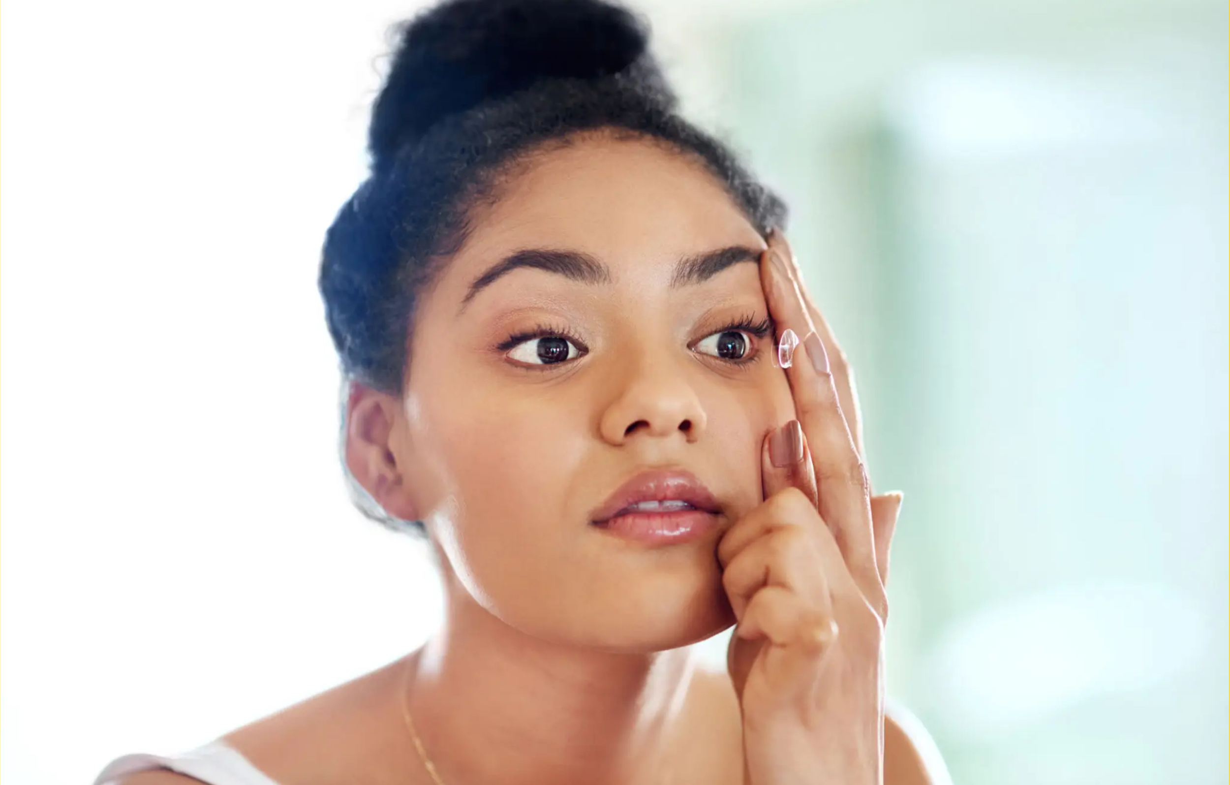 A woman with dark hair styled in a bun gently touches her temple with her fingers, looking slightly to her right with a surprised or curious expression.