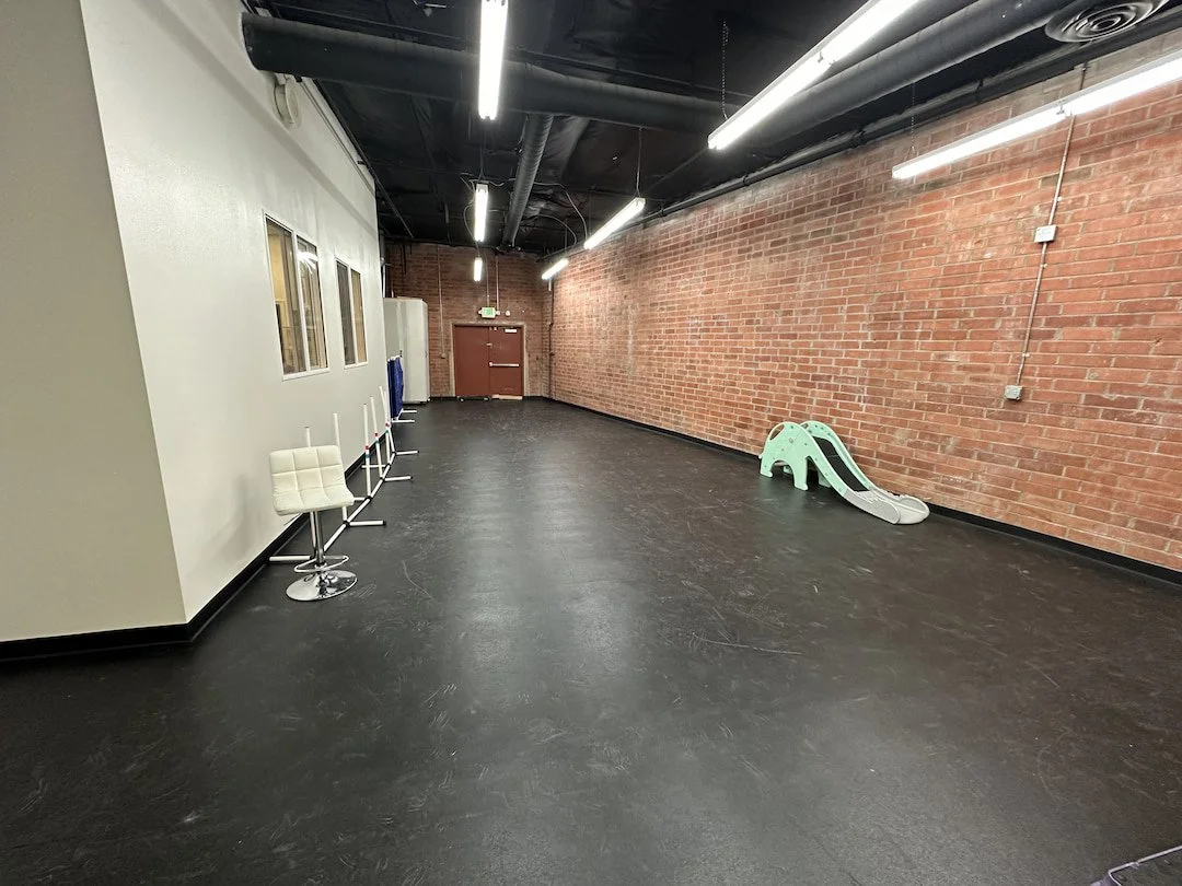 Empty indoor space with black flooring, brick wall on the right, white wall with three windows on the left, white chairs stacked on the left, children's slide on the right, ceiling with exposed black pipes and long fluorescent lights.