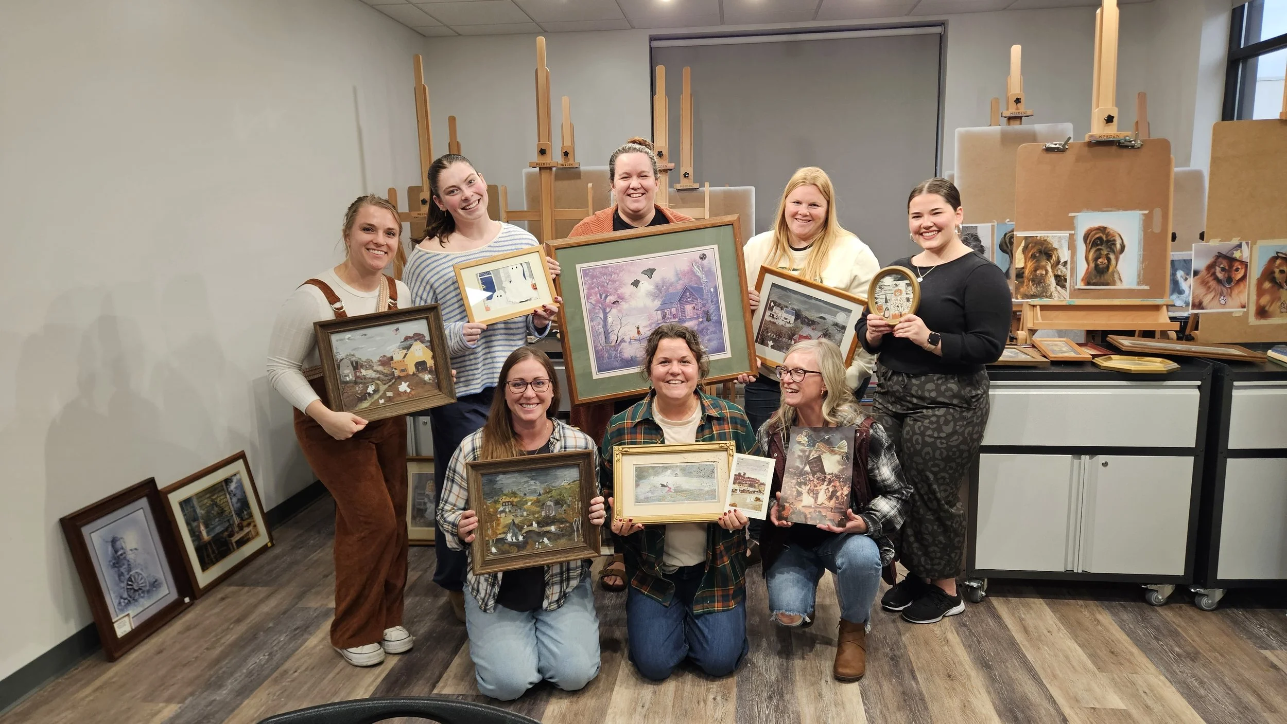 Group of women in an art classroom holding framed paintings and photographs of landscapes and animals, with easels and more artwork in the background.