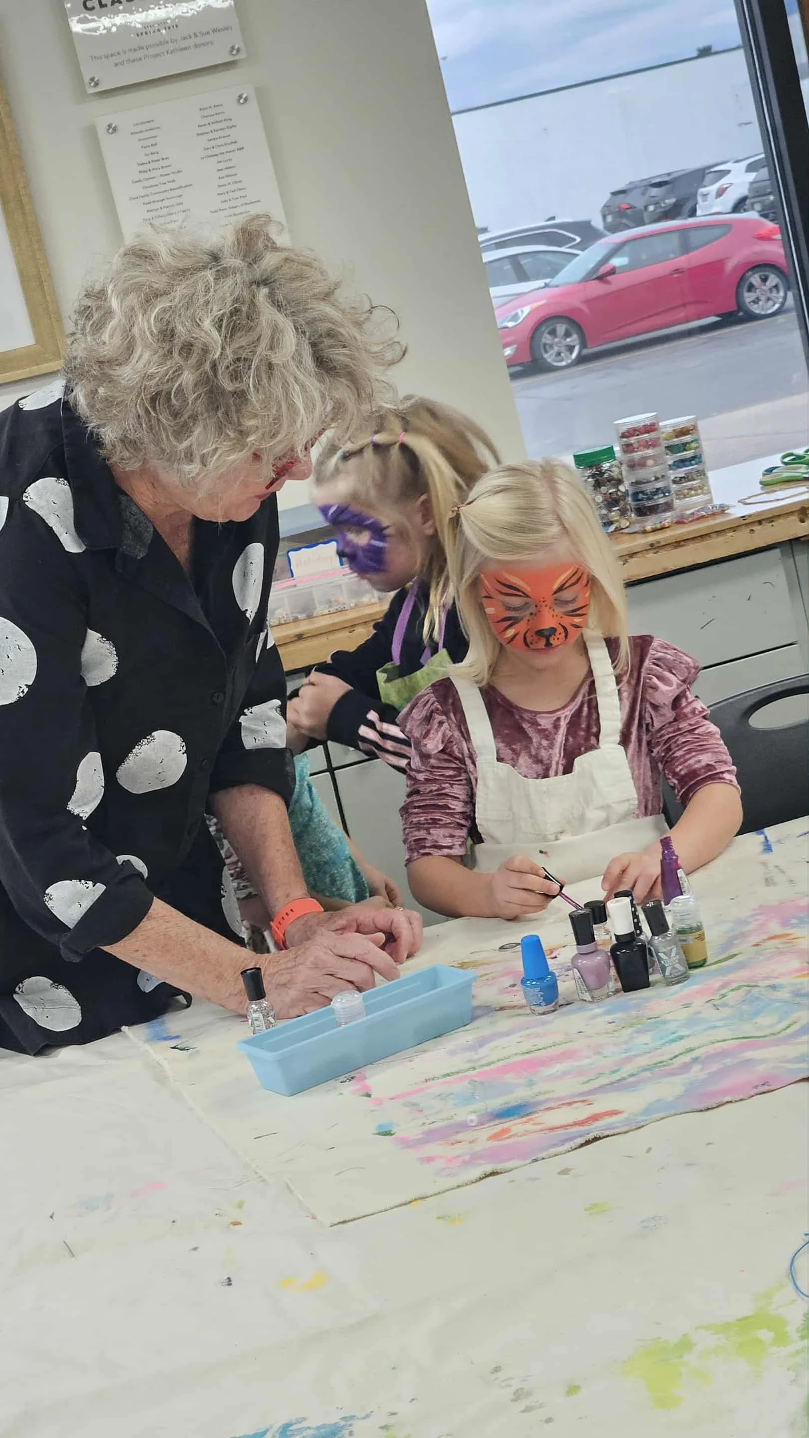 An elderly woman with curly gray hair helps two young girls with face painting at a table. The girls have their faces painted like a tiger and a zebra, and their faces are painted with vibrant colors. The scene takes place indoors with a window showing parked cars outside.