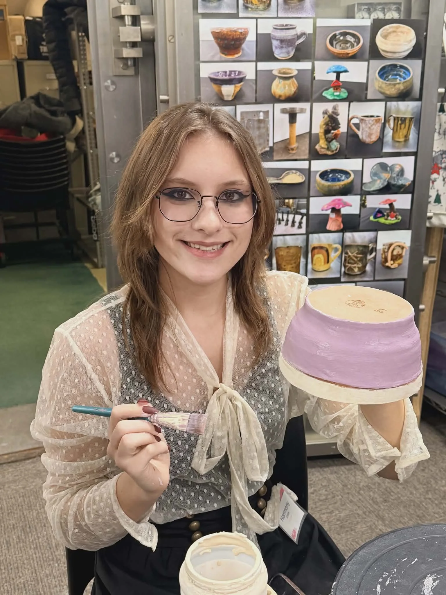 A woman in glasses holding a paintbrush and a painted bowl in Olio's pottery studio, with a background of shelves and pictures of various ceramic or pottery items.