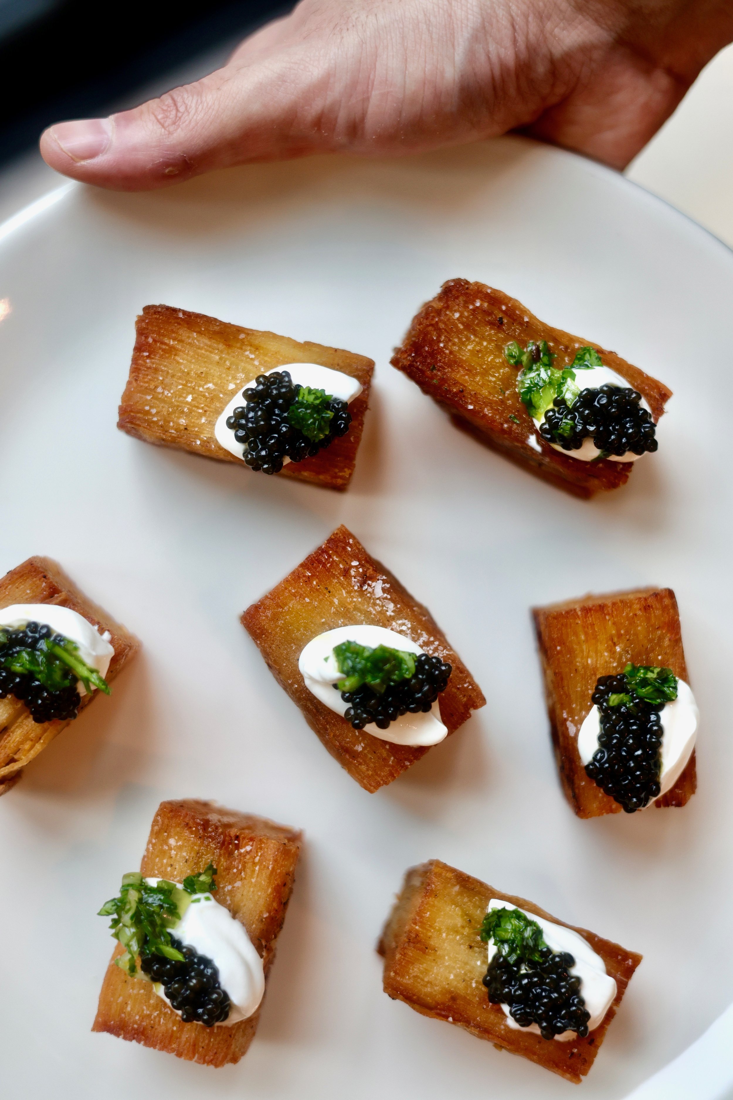 Close-up view of multiple rectangular pieces of fried bread topped with dollops of sour cream, black caviar, and chopped chives, arranged on a white plate.