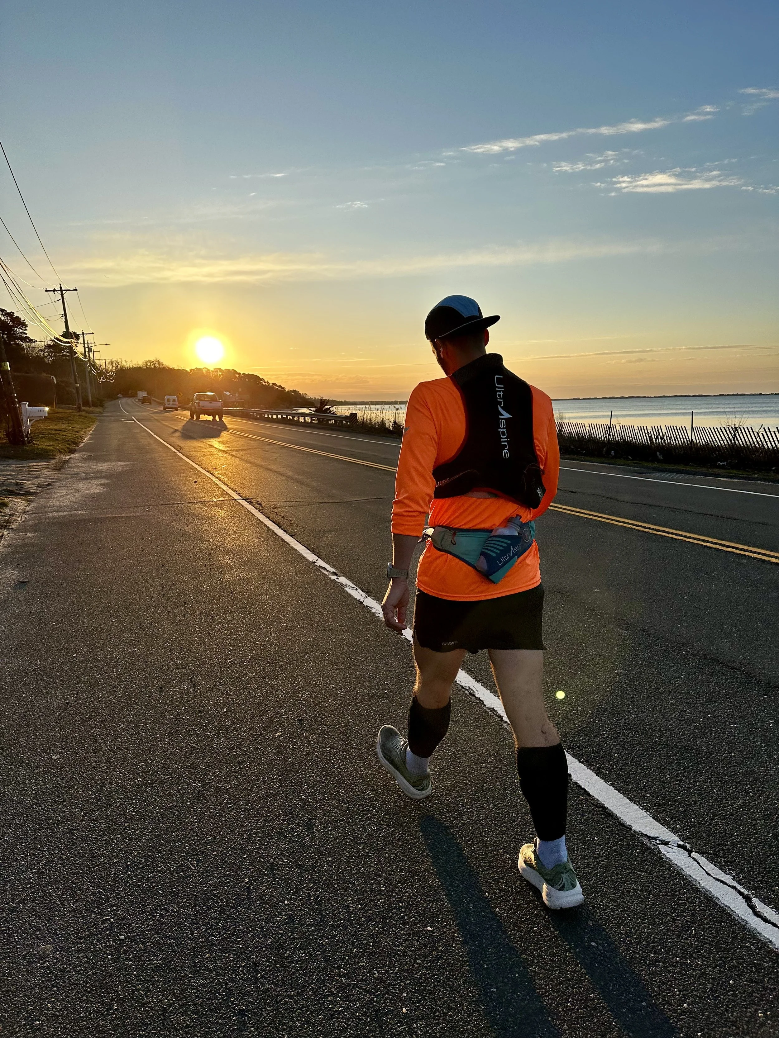 Man dressed in athletic gear running on a road during sunset near water with cars in the distance.
