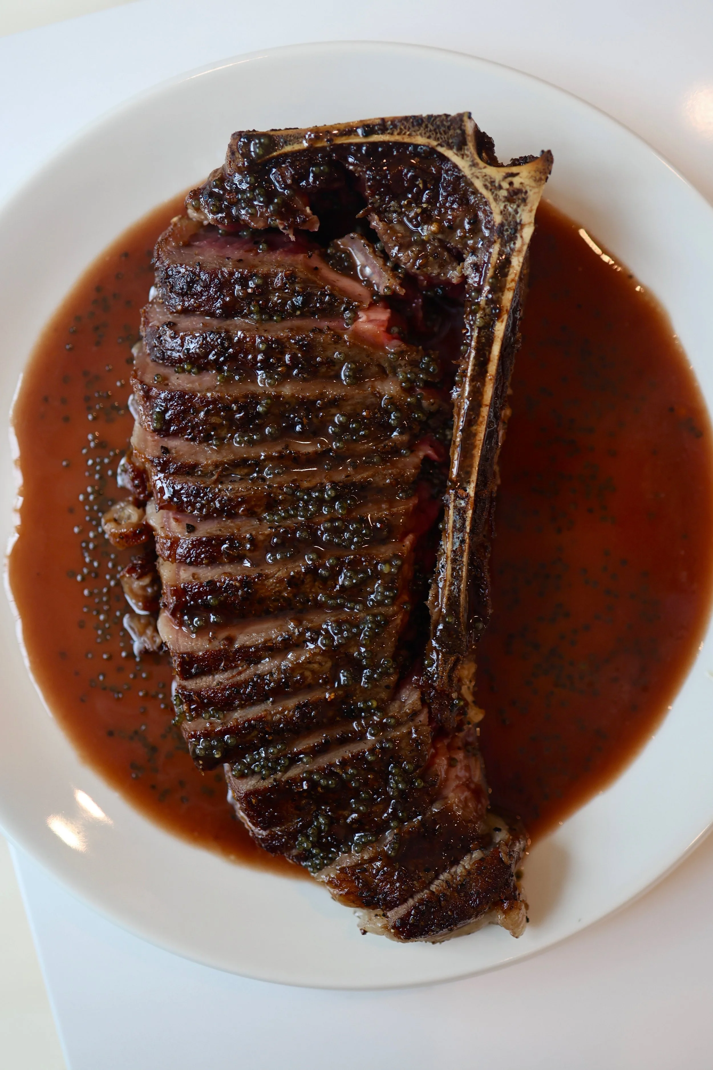 A cooked bone-in steak topped with peppercorn sauce on a white plate.
