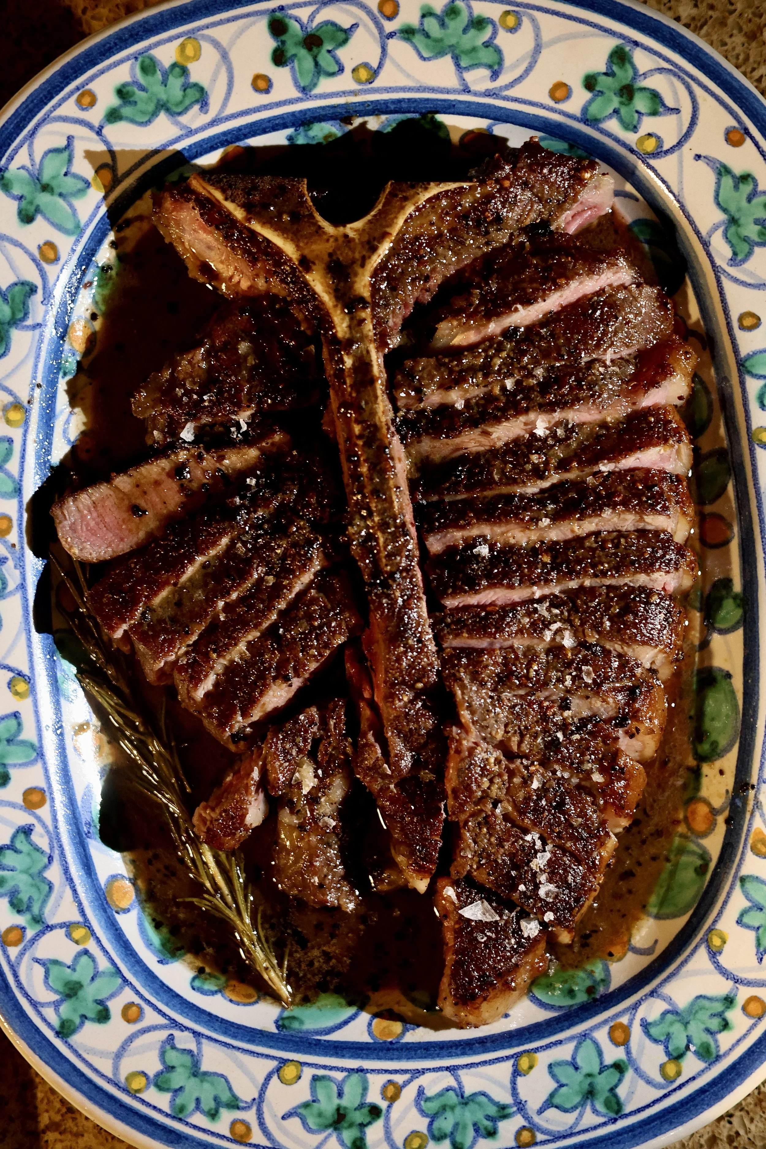 Cooked slices of seasoned and grilled steak on a decorative ceramic plate with salt sprinkled on top and a sprig of rosemary.