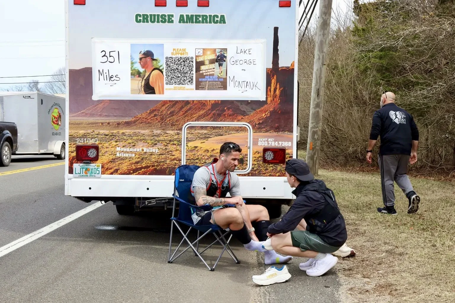 A man receiving a foot treatment from another man beside a promotional truck for a charity event on a rural road, with another man walking away in the background.
