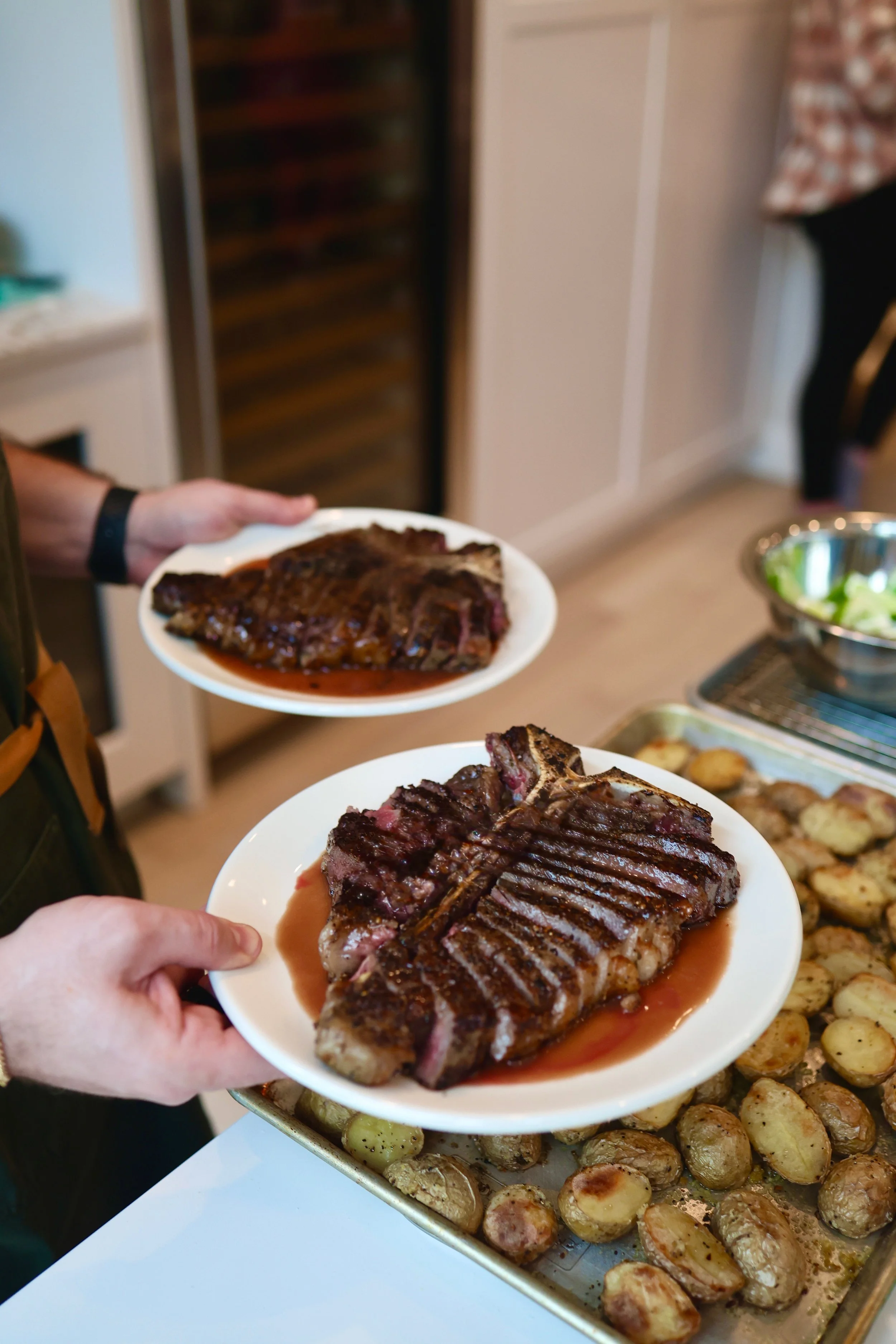Hands holding plates with grilled steaks, beef, and vegetables, served at a buffet-style setting with roasted potatoes in a metal tray.