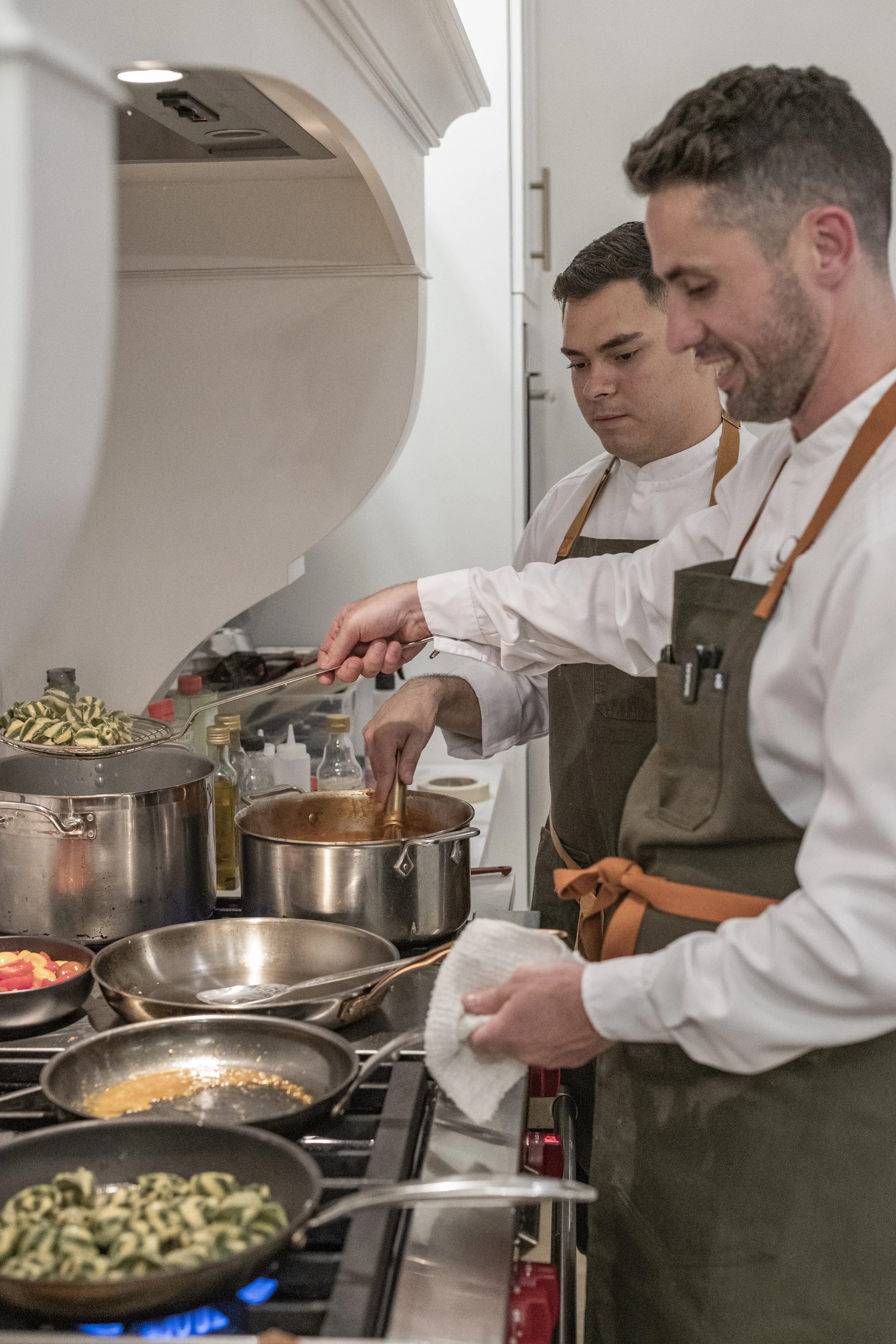 Two male chefs are cooking at a kitchen stove, stirring pots and preparing food, wearing white coats and aprons.