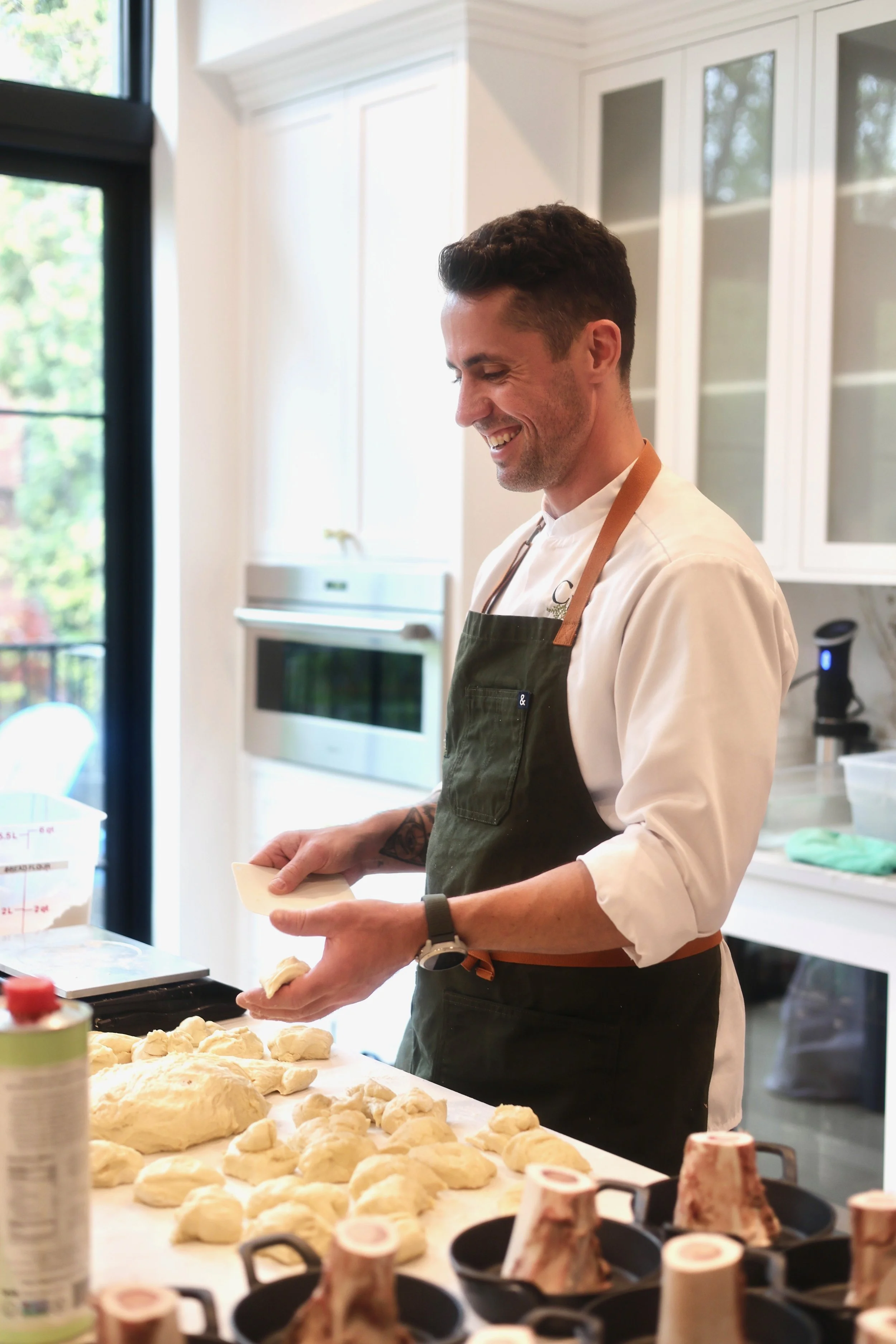 Man with dark hair smiling and wearing a white shirt, black apron, and wristwatch, working in a bright kitchen with white cabinets and large window, holding a piece of dough. The table in front has dough, and there are various baking ingredients and 