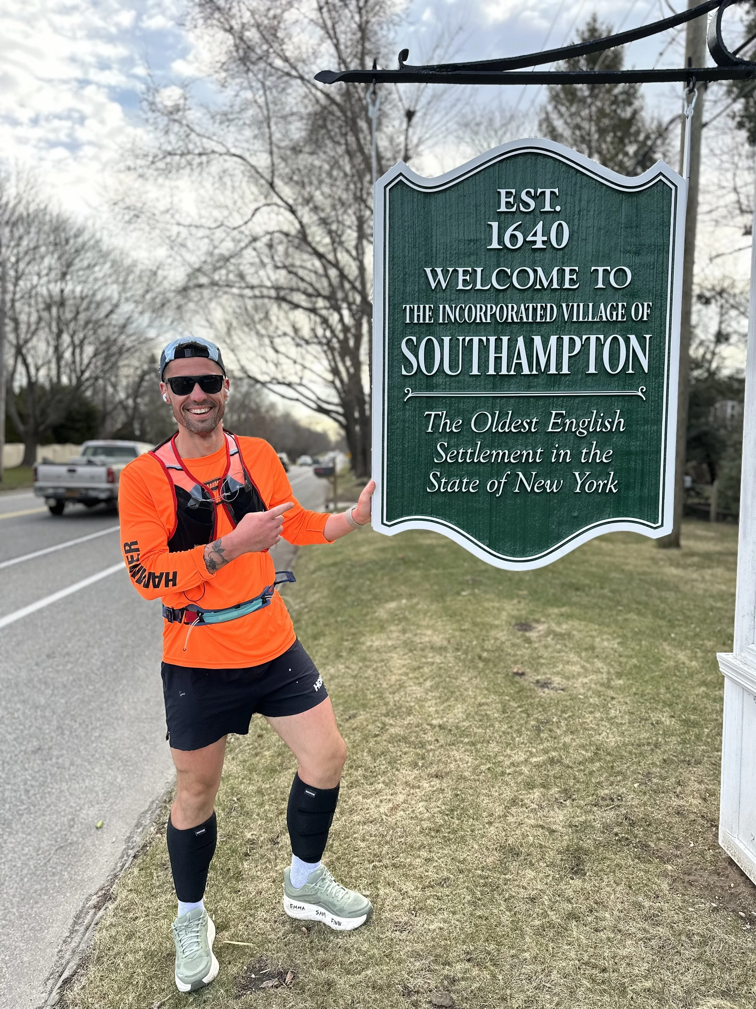 Man in orange athletic shirt and black shorts standing next to a green welcome sign for Southampton, New York, pointing at the sign with his right hand and smiling. The sign indicates Southampton was established in 1640 and is the oldest English sett