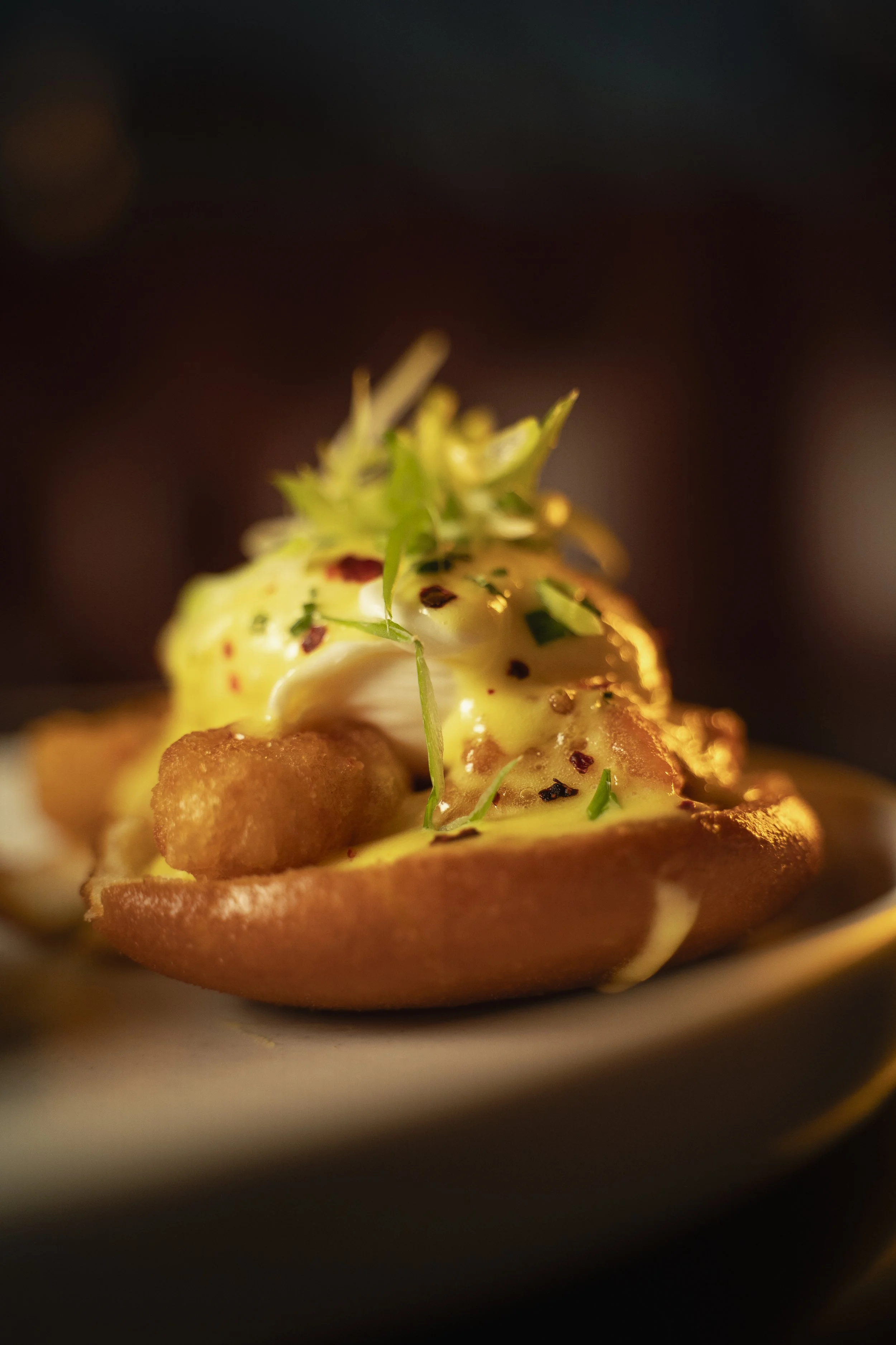 Close-up of a gourmet dish featuring a poached egg on toast, topped with hollandaise sauce, chopped herbs, and seasoning, served on a plate.