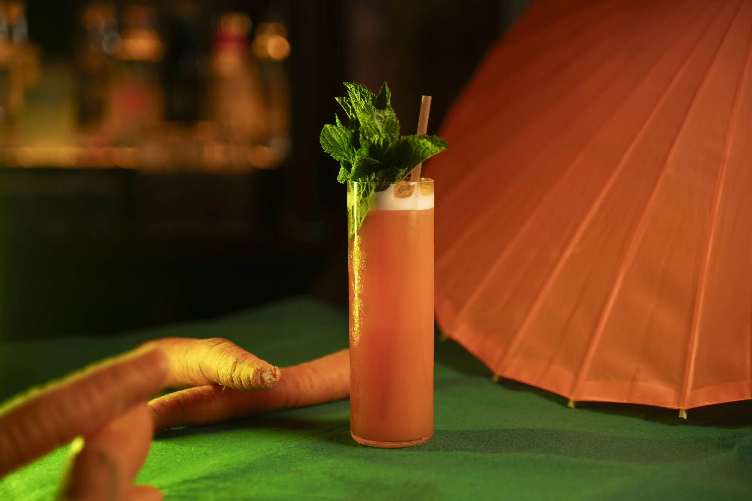 A pink cocktail garnished with mint leaves, served in a tall glass with a straw, set against a vibrant green surface with an orange parasol in the background.
