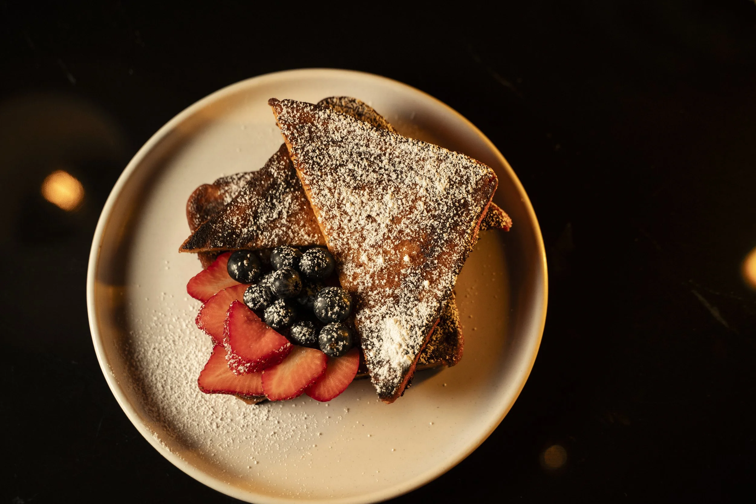A dessert plate with French toast dusted with powdered sugar, garnished with sliced strawberries and blueberries, on a white dish against a dark background.