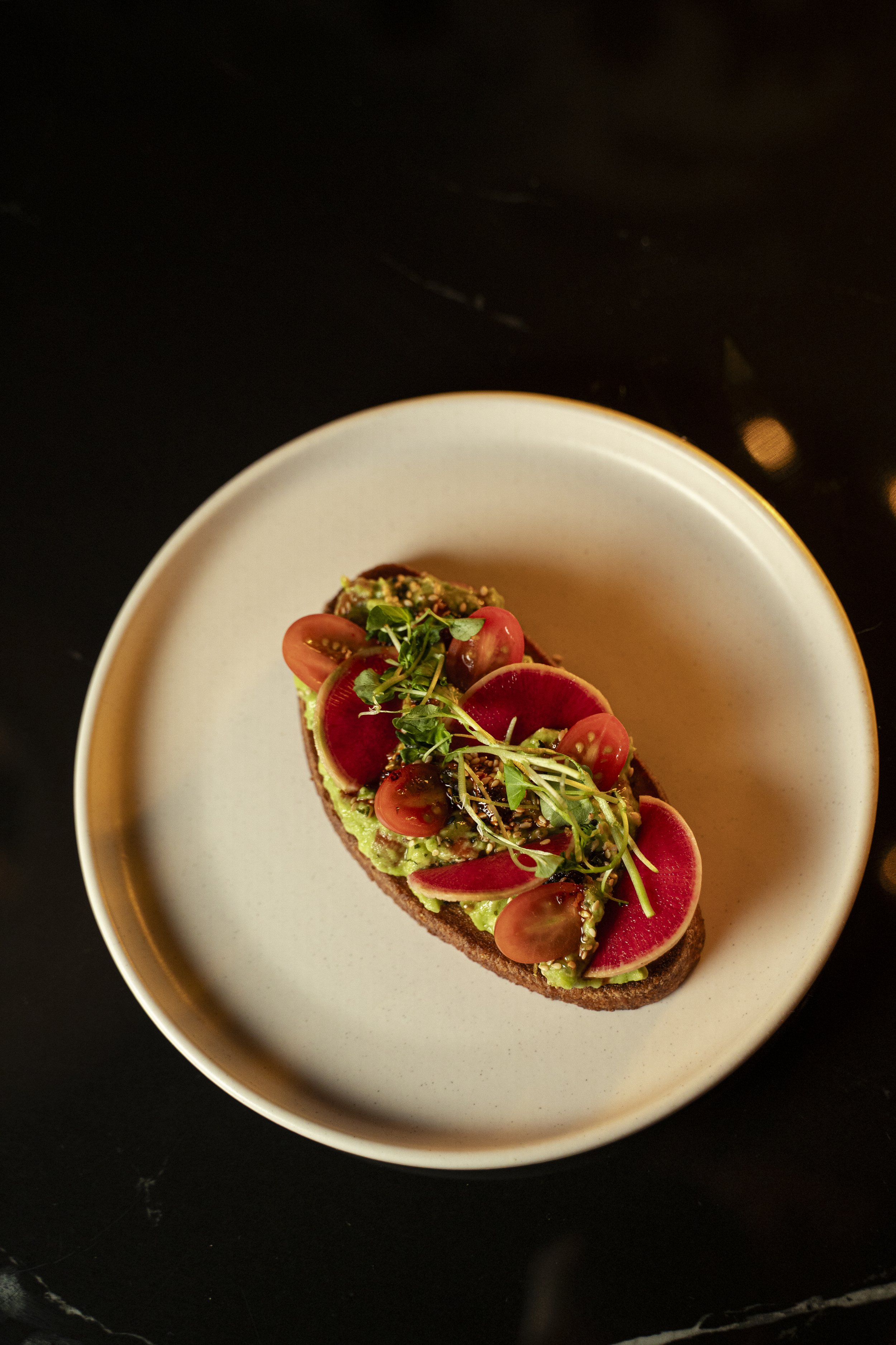 Open-faced sandwich with sliced cherry tomatoes, radish slices, microgreens, and guacamole on toasted bread, served on a white plate.
