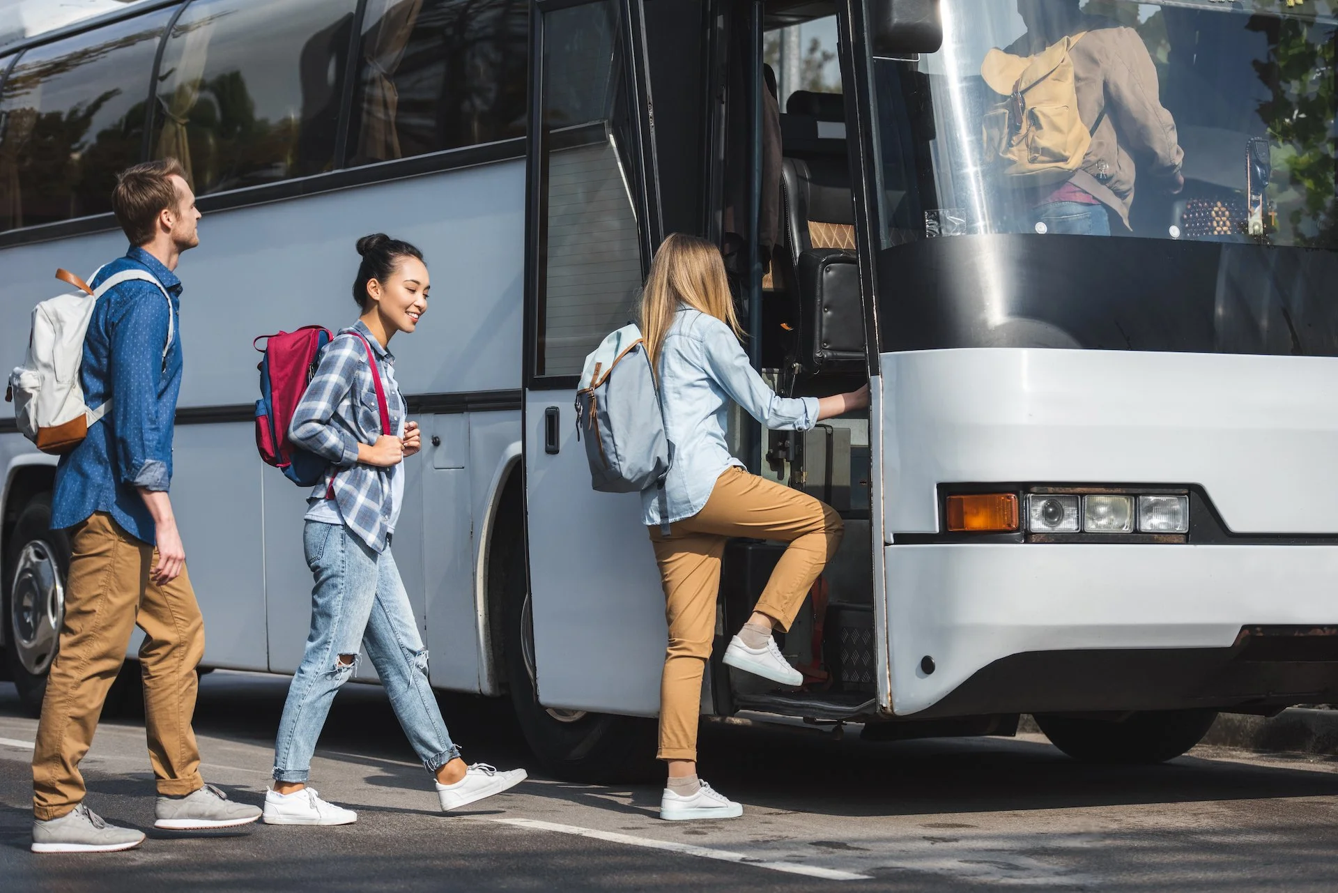 Three young adults boarding a tour bus, two women and one man, carrying backpacks, dressed casually, with one woman entering the bus and the others walking on the street.