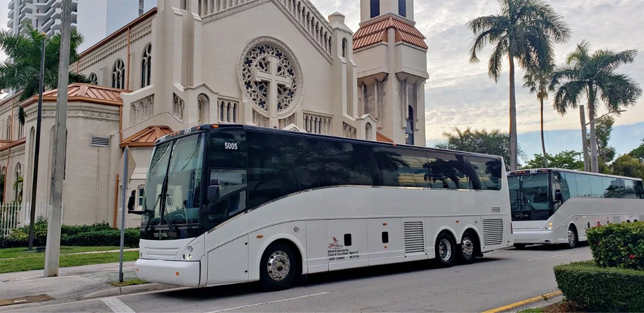 Two white tour buses parked on the street in front of a church with tall palm trees and a cloudy sky in the background.