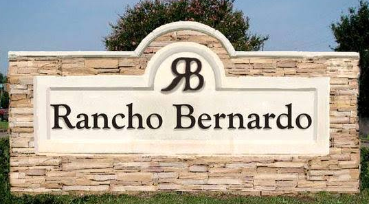 Sign with the text "Rancho Bernardo" and a logo with the letters "RB" on a brick background, outdoors with trees in the background.