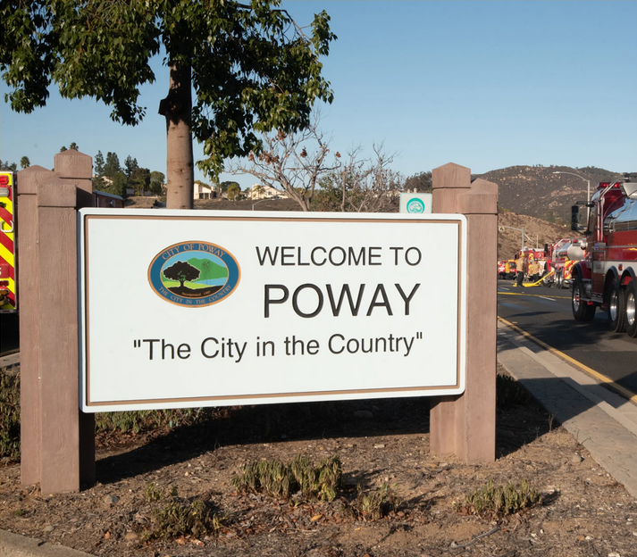 Welcome sign for Poway, California, with the city logo, slogan 'The City in the Country,' and emergency vehicles in the background.