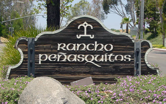 Wooden sign for Rancho Peñasquitos with a decorative symbol above the name, surrounded by purple flowers and rocks.