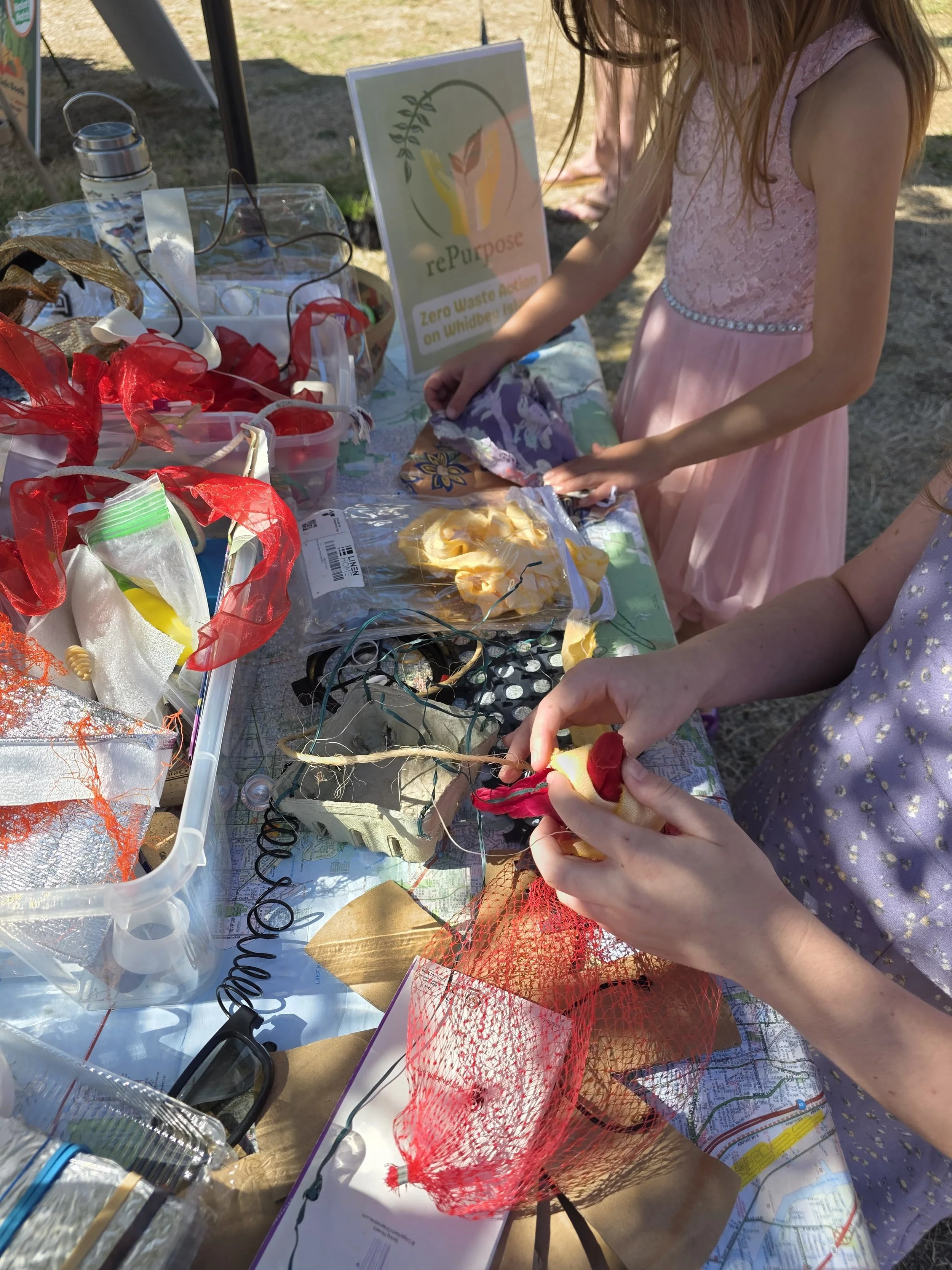 A table with lots of repurposed arts and crafts materials strewn about. Two young, caucasian children play with some ribbons and fabric.