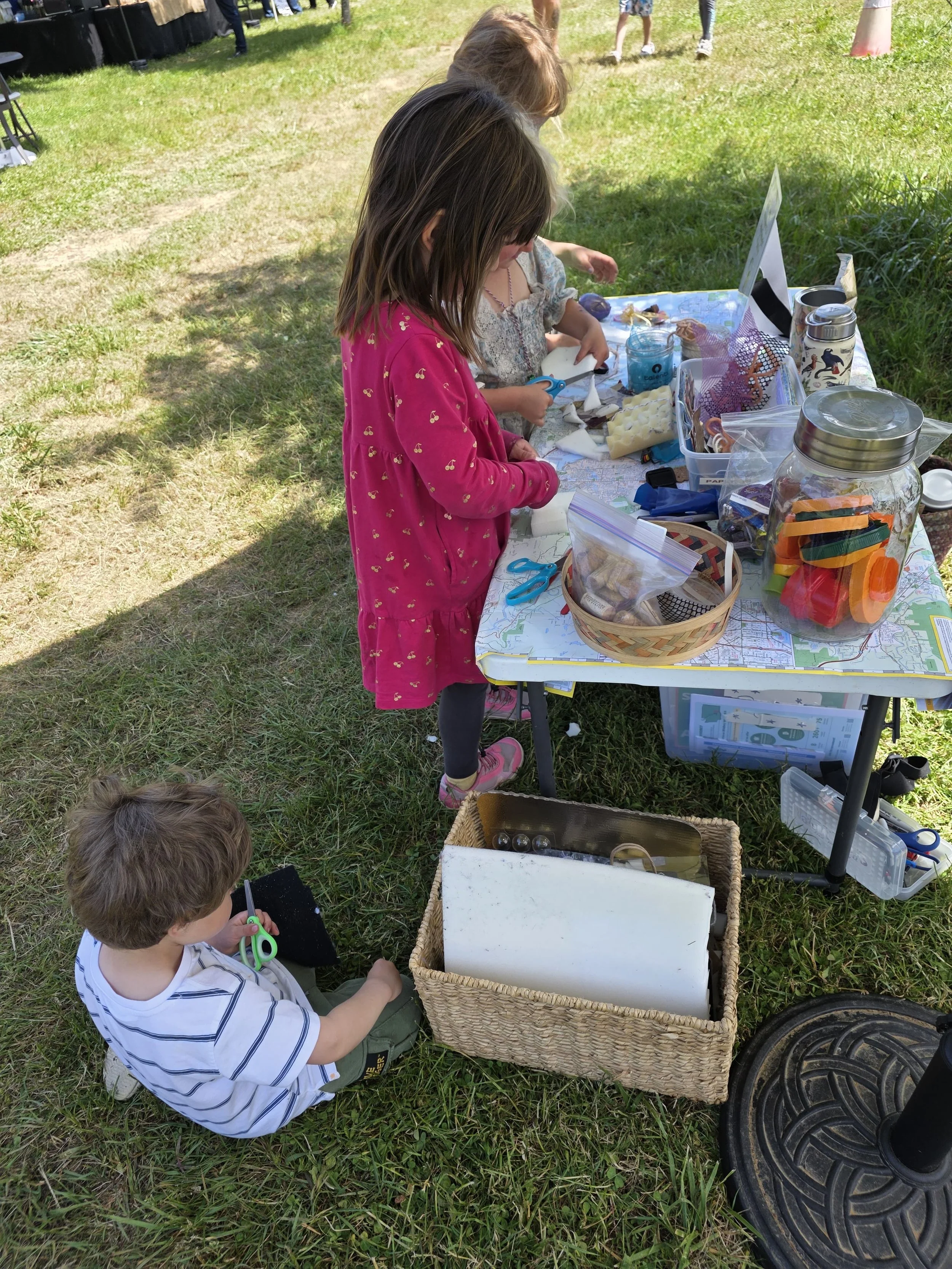 Three young caucasian children stand at a table and play with repurposed arts and crafts materials, including corks, paper scraps, and ribbons.