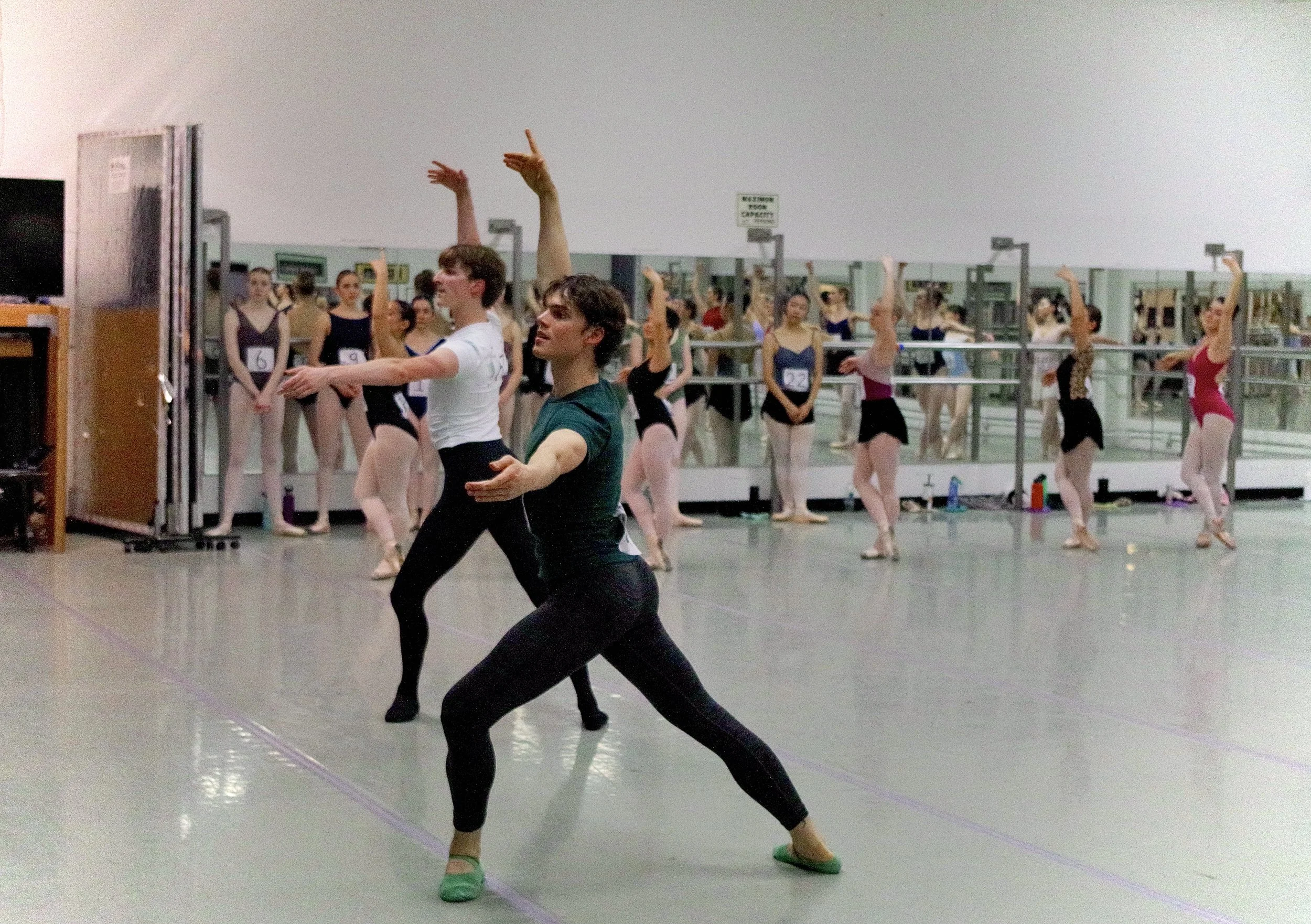 Ballet dancers practicing in a dance studio with two instructors leading and mirrors lining the wall.