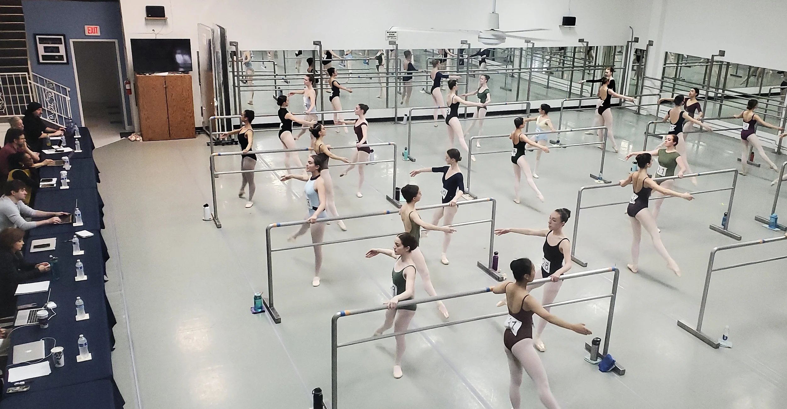 Ballet dancers practicing at ballet barres in a dance studio, with judges observing and taking notes at a table on the left.