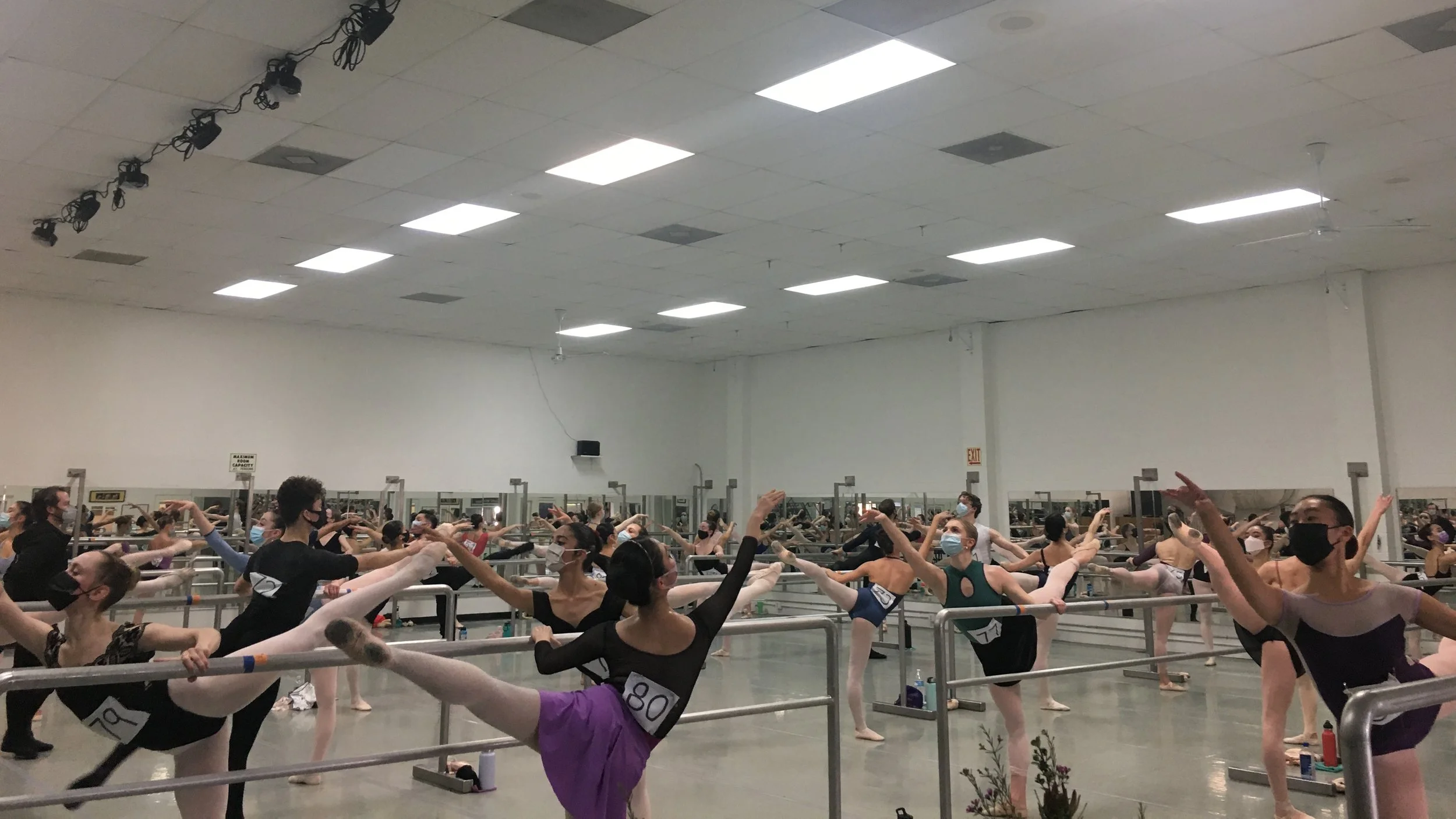 Ballet dancers in a dance studio practicing at ballet barre, wearing masks and numbered bibs.