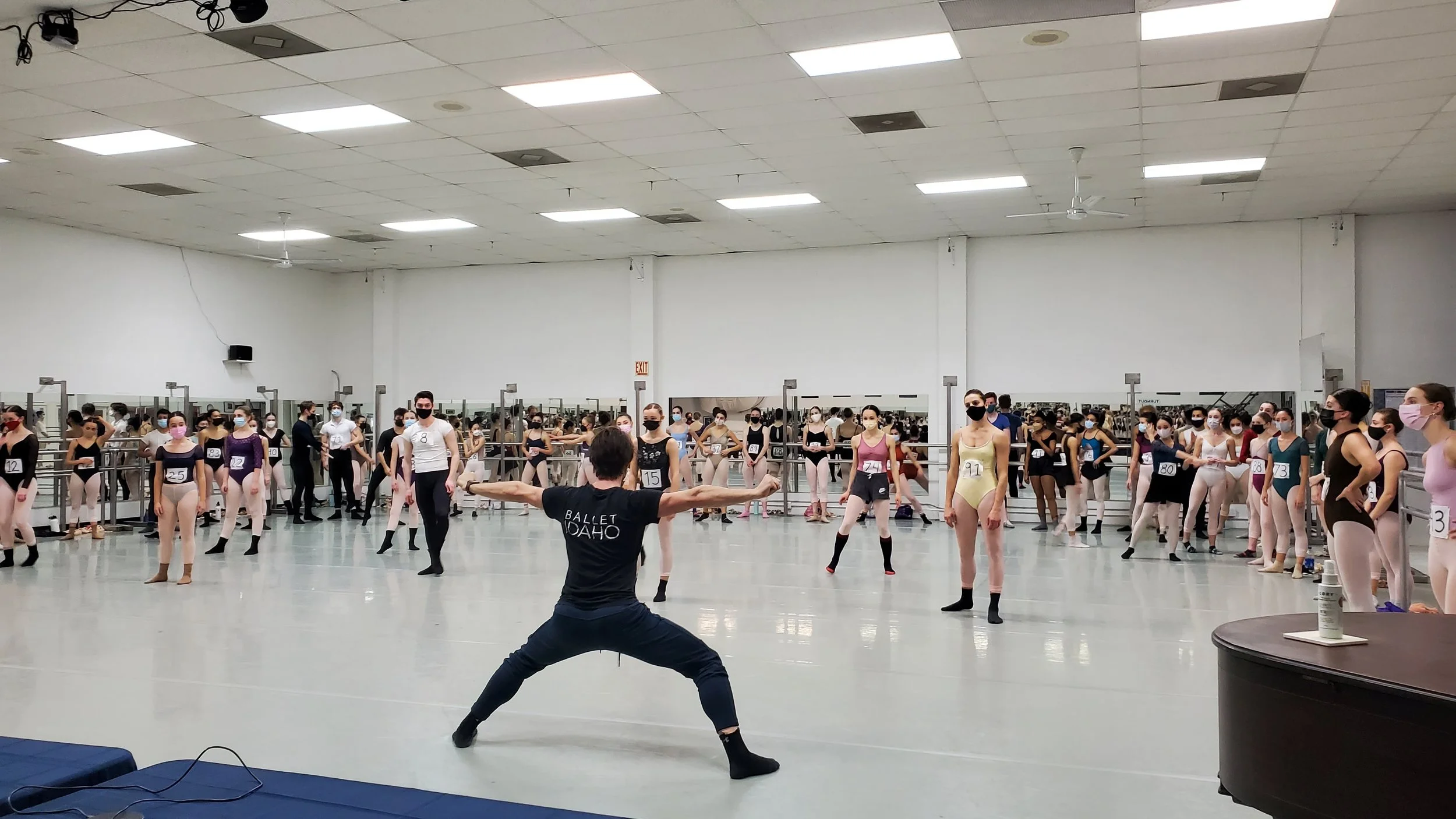 Ballet dancers practicing in a studio with an instructor leading them, all wearing masks and numbered costumes.